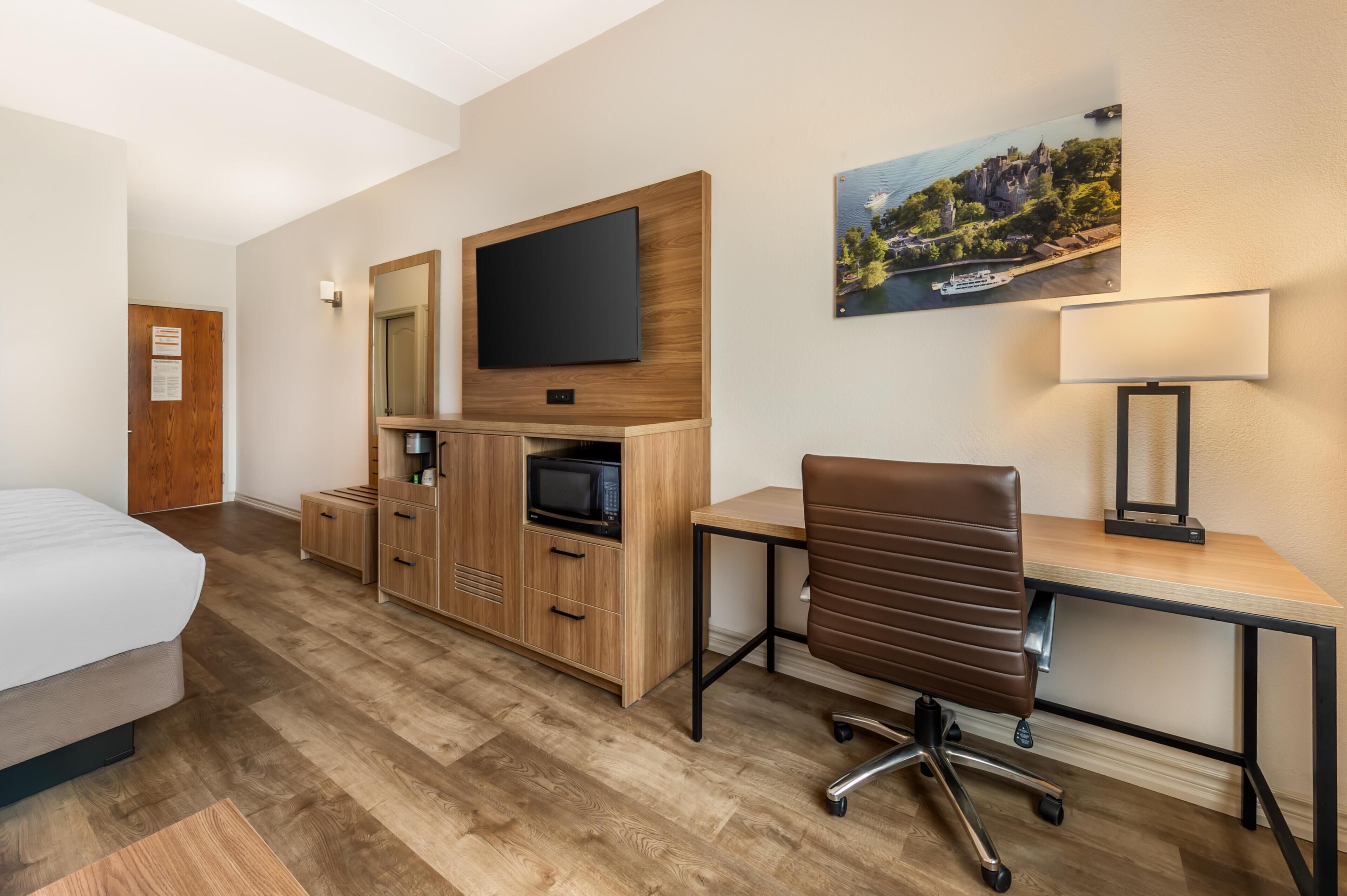 A hotel room with a bed, a desk with a chair, a television, a microwave, and a painting on the wall.