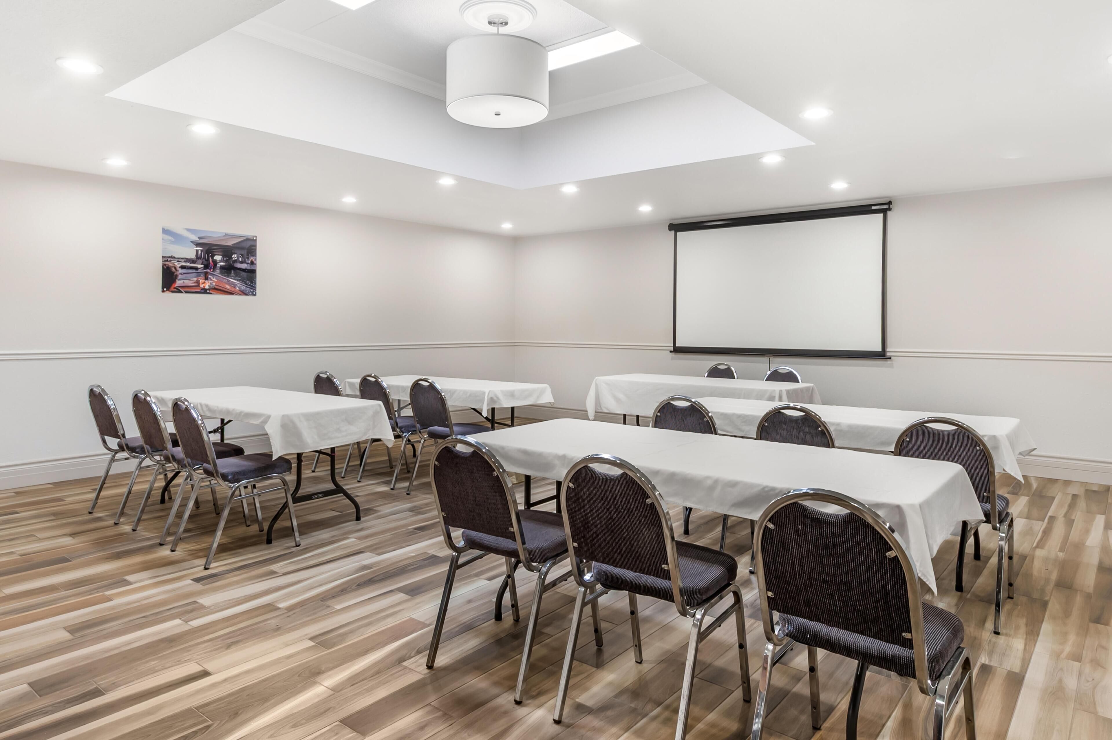 A conference room with white tables, dark chairs, a projector screen, and a ceiling light.