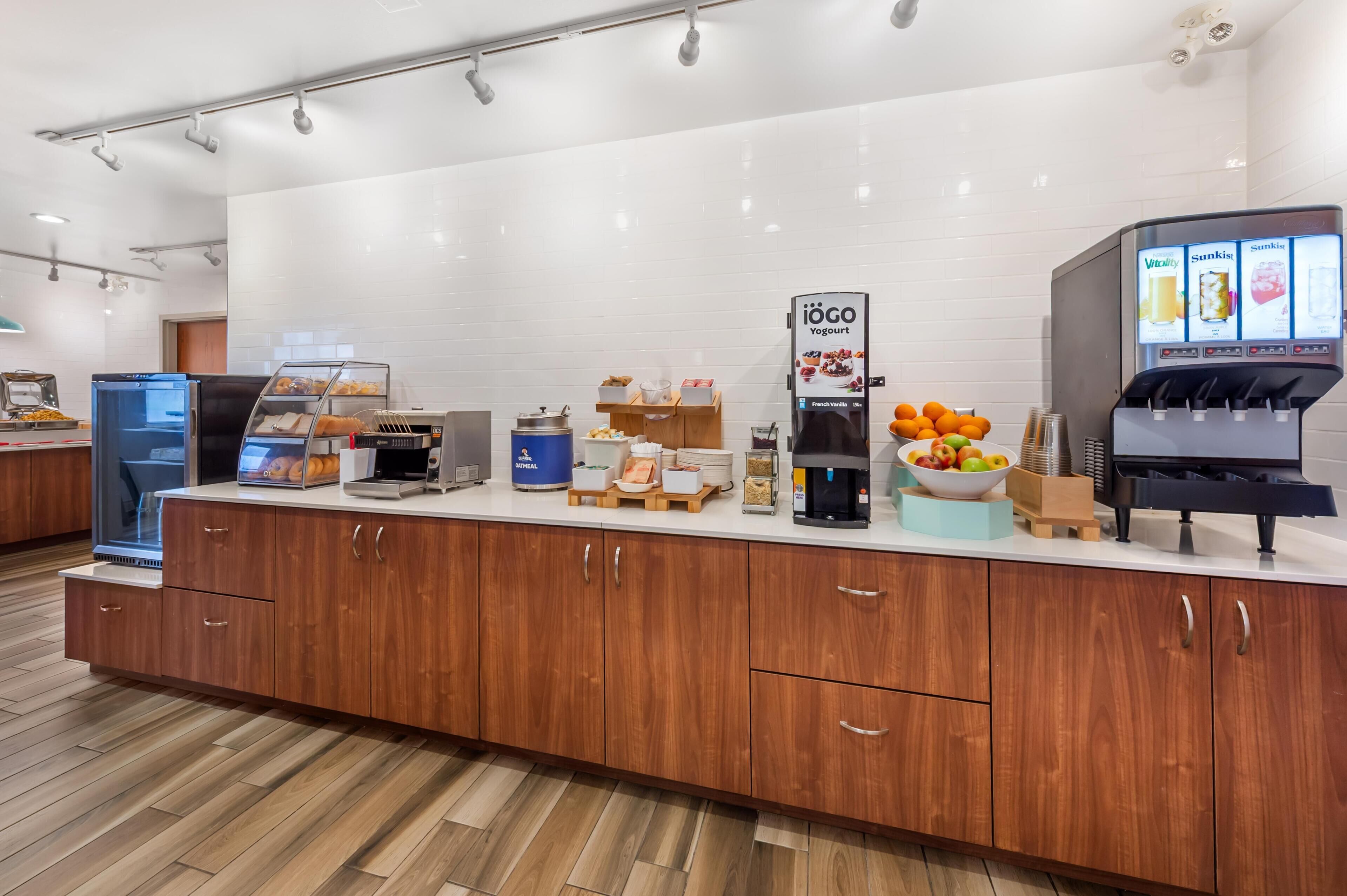 Zona de desayuno autoservicio con dispensador de yogur, máquina de café y diversas opciones de alimentos.