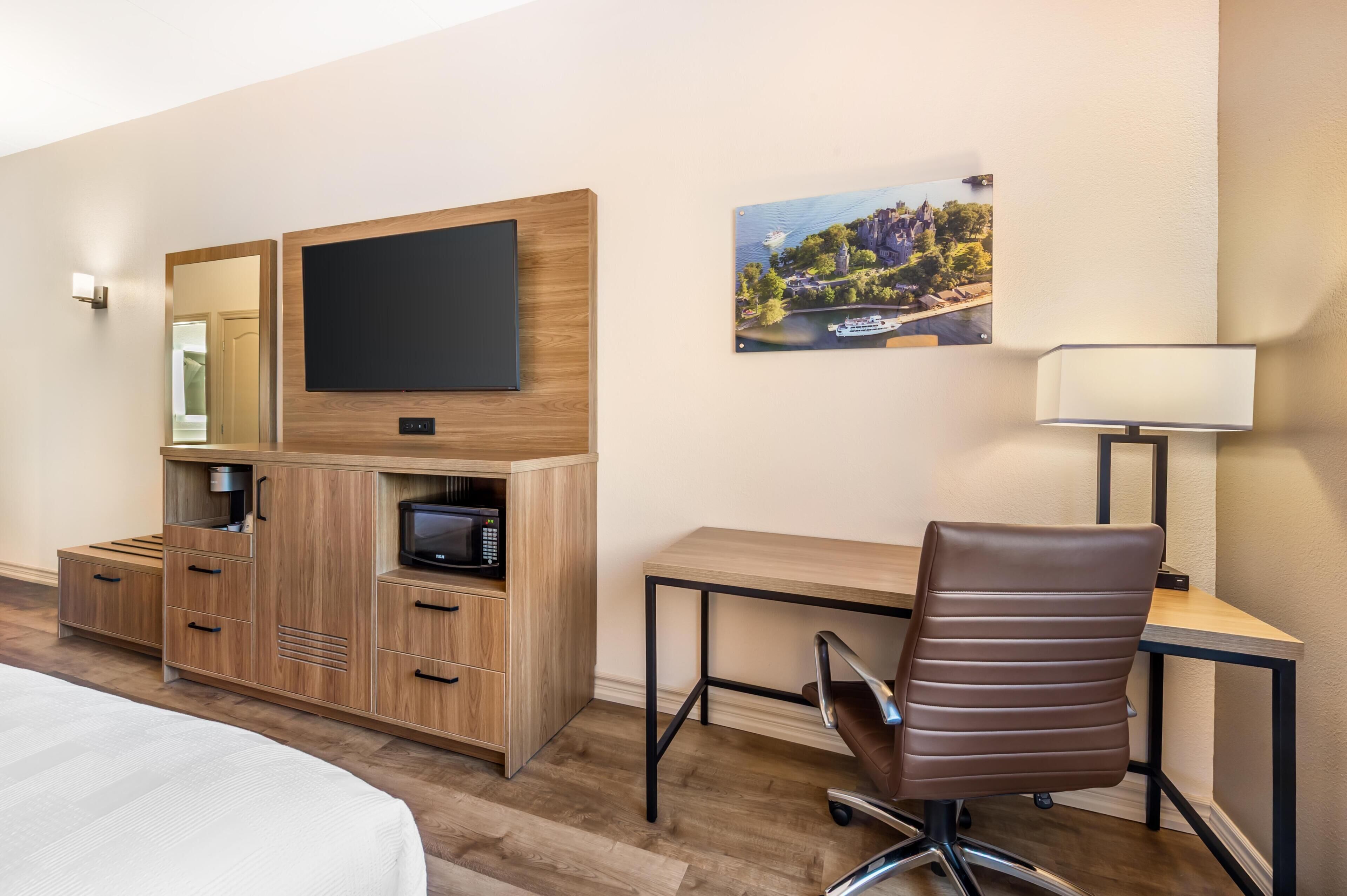 A hotel room with a bed, a wooden desk with a chair, a television, a microwave, a lamp, and a framed picture on the wall.