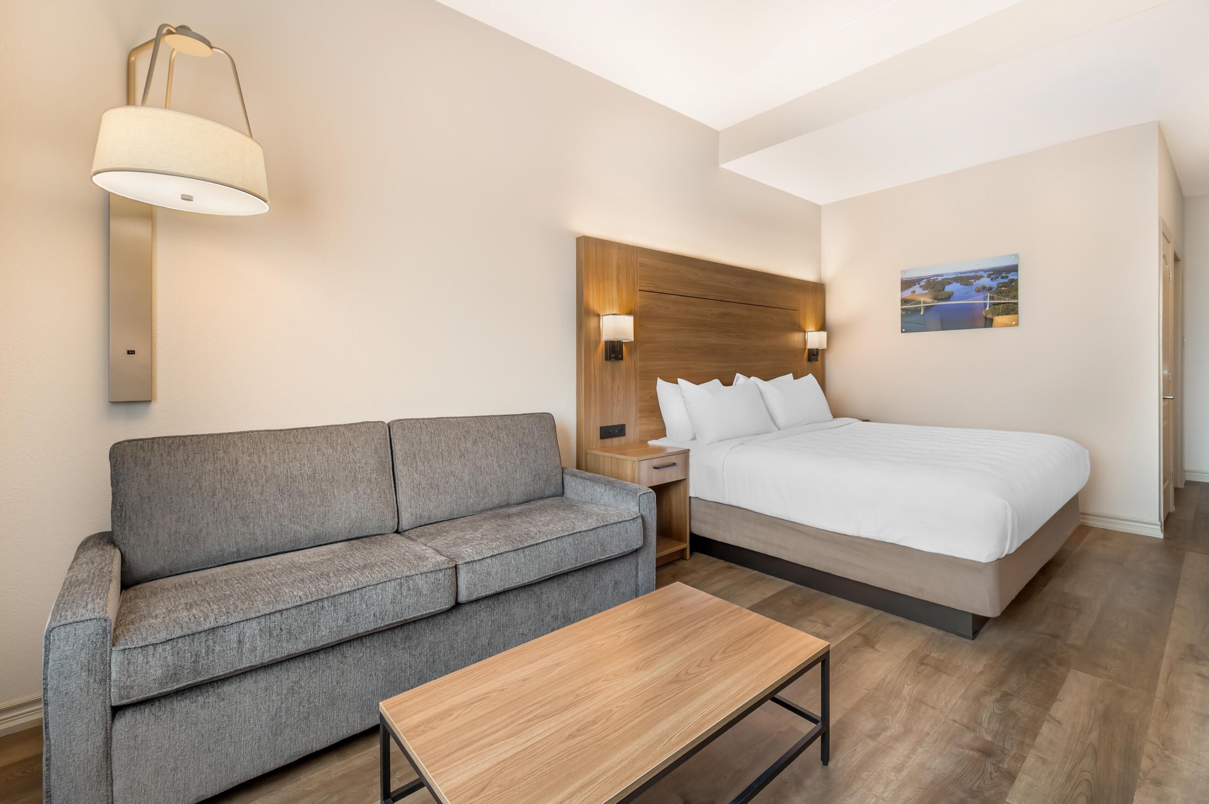 Habitación de hotel con una cama, un sofá gris, una mesa de madera, una mesita de noche y una lámpara de pared.