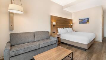 Habitación de hotel con una cama, un sofá gris, una mesa de madera, una mesita de noche y una lámpara de pared.