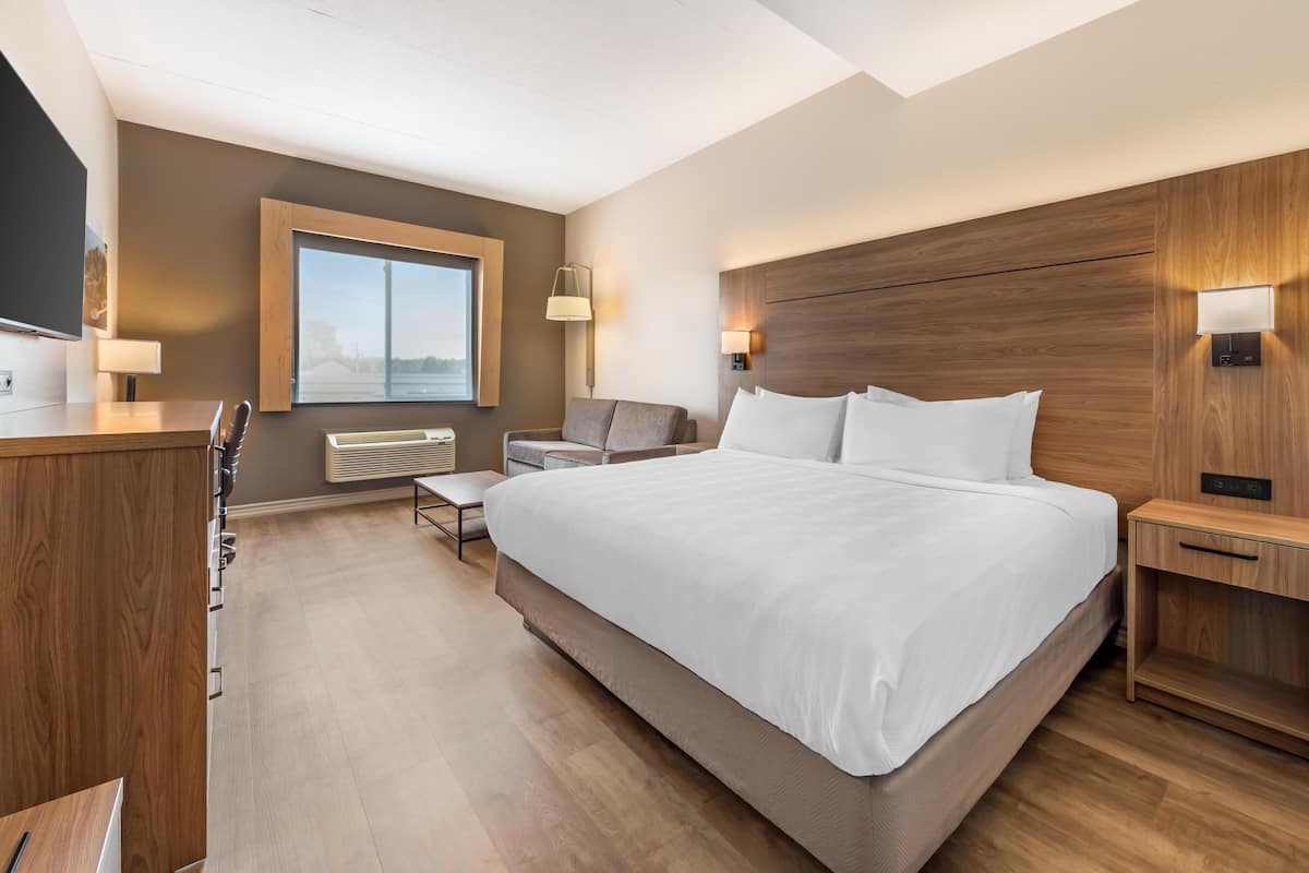 A modern hotel room with a large bed, a desk, a chair, a bench, and a window with a view.