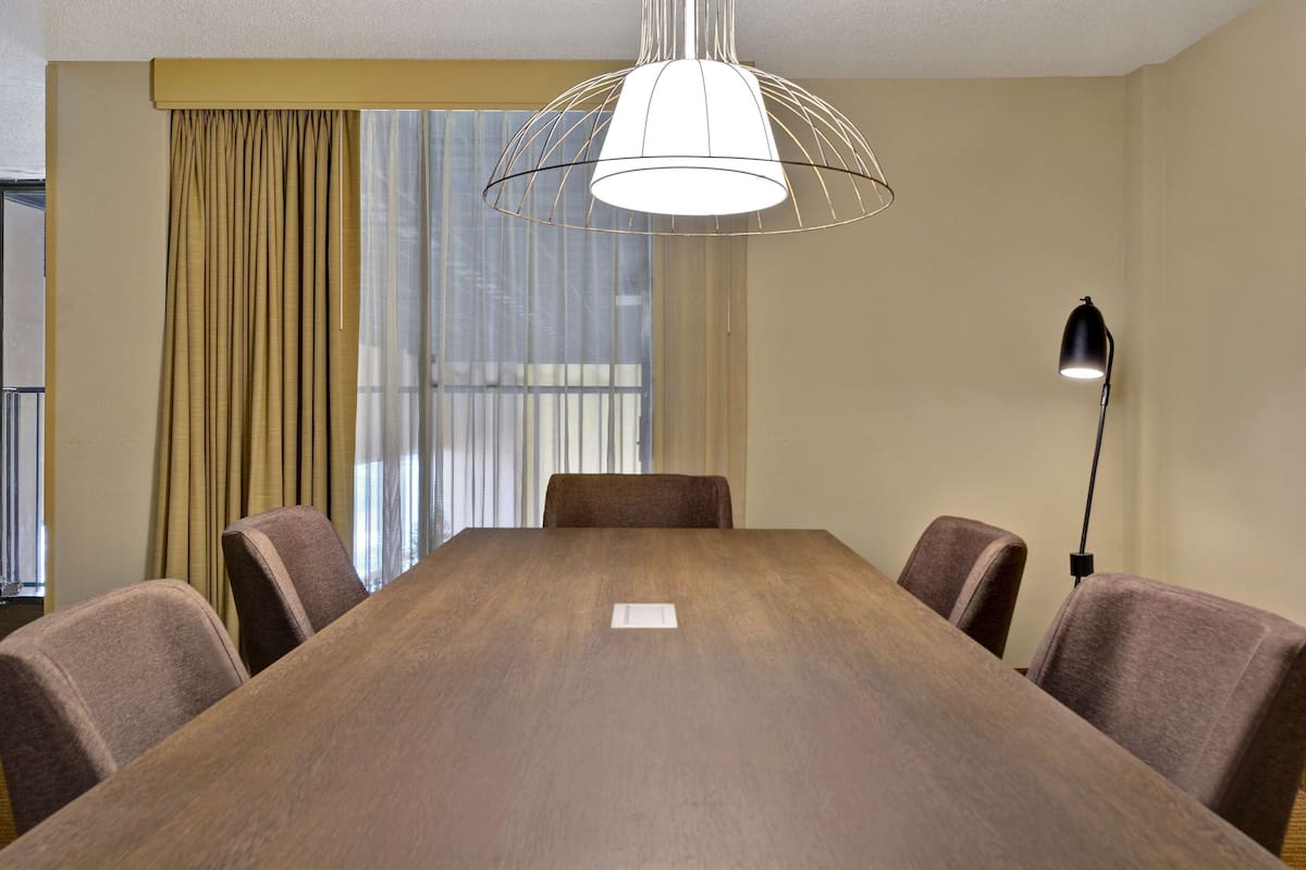 A conference room with a large wooden table, four chairs, a hanging light fixture, a floor lamp, and a window with curtains.