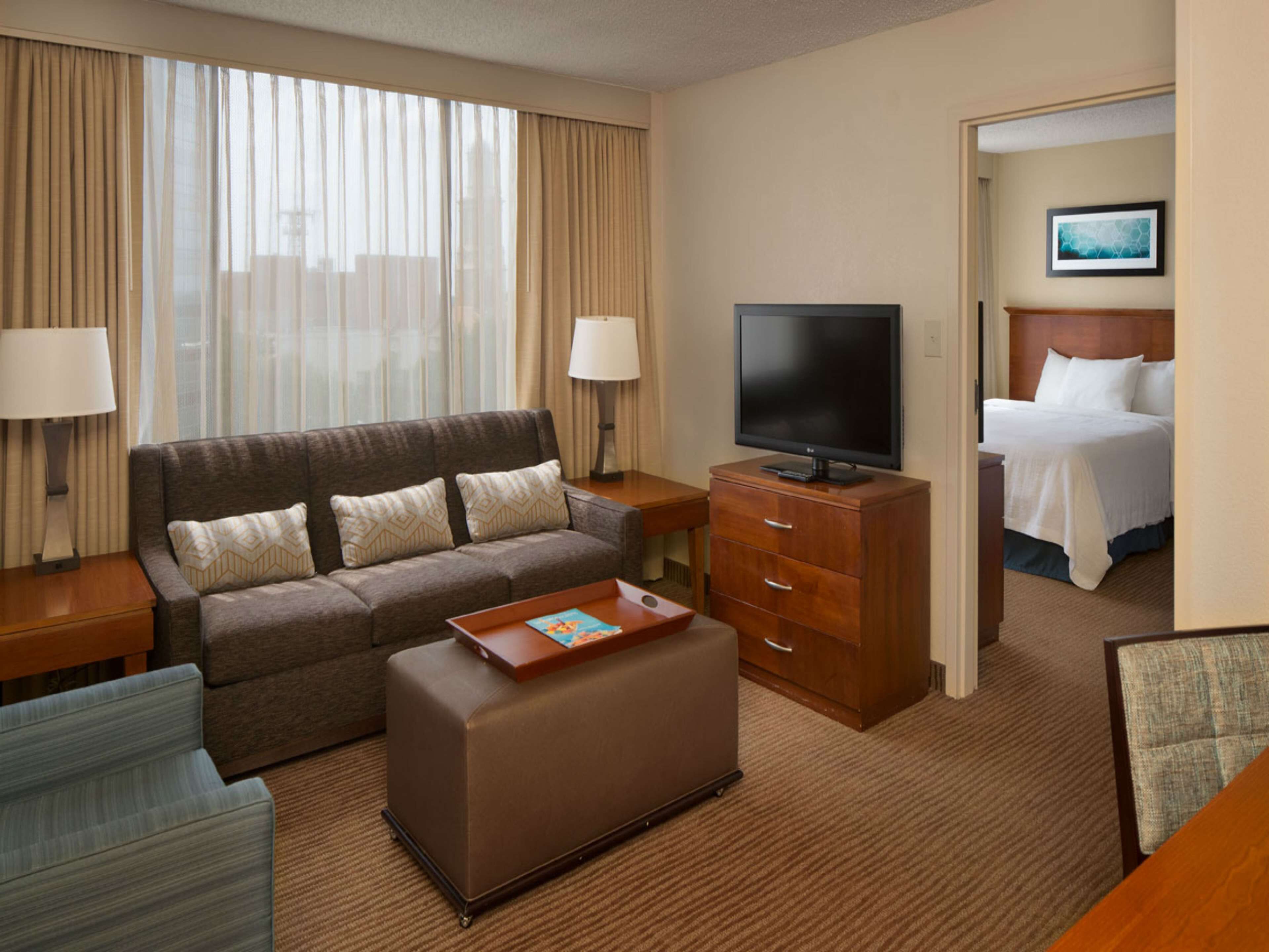 A hotel room with a sofa, ottoman, TV, and a bed with a headboard.