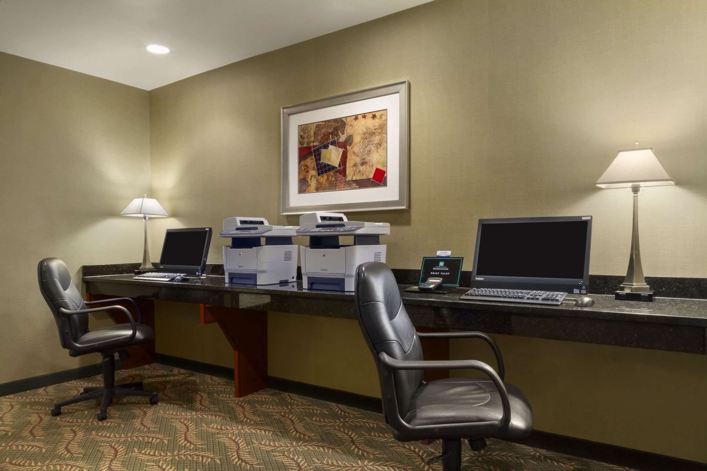 A hotel reception desk with two computer stations, two chairs, a framed artwork on the wall, and two lamps.
