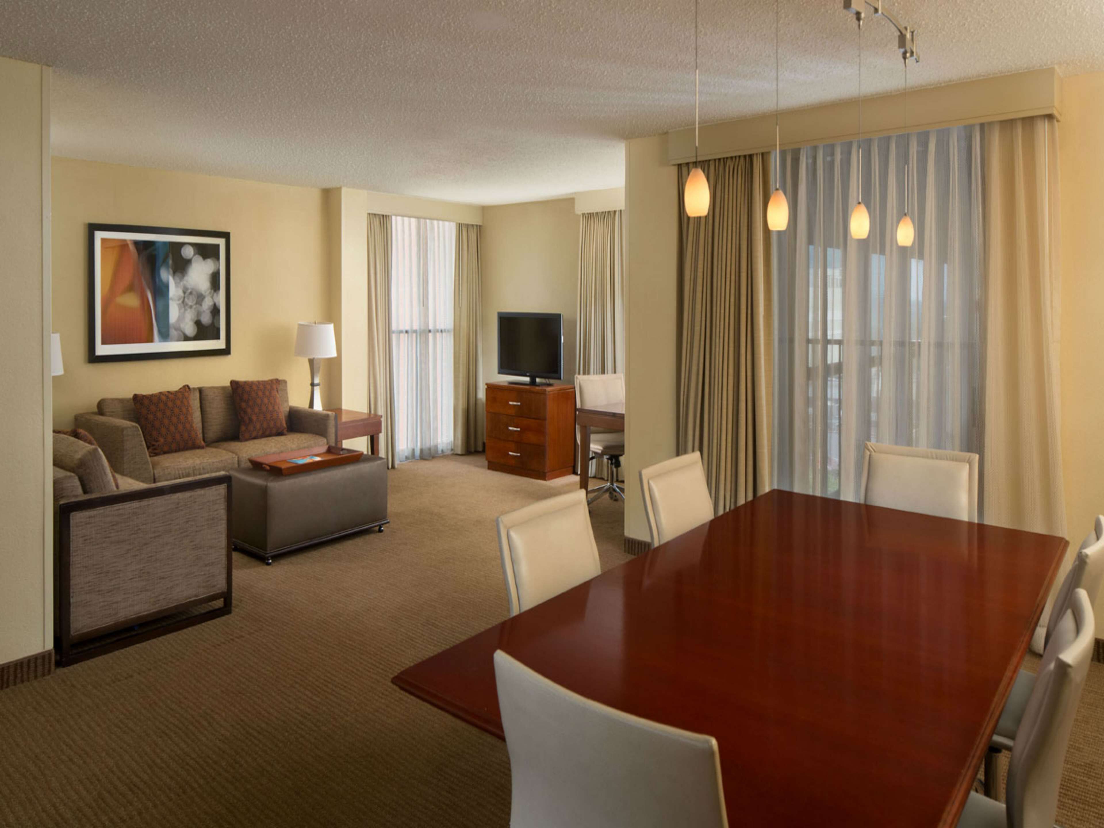 A hotel room with a living area, a dining table with chairs, a TV, and a large window with curtains.
