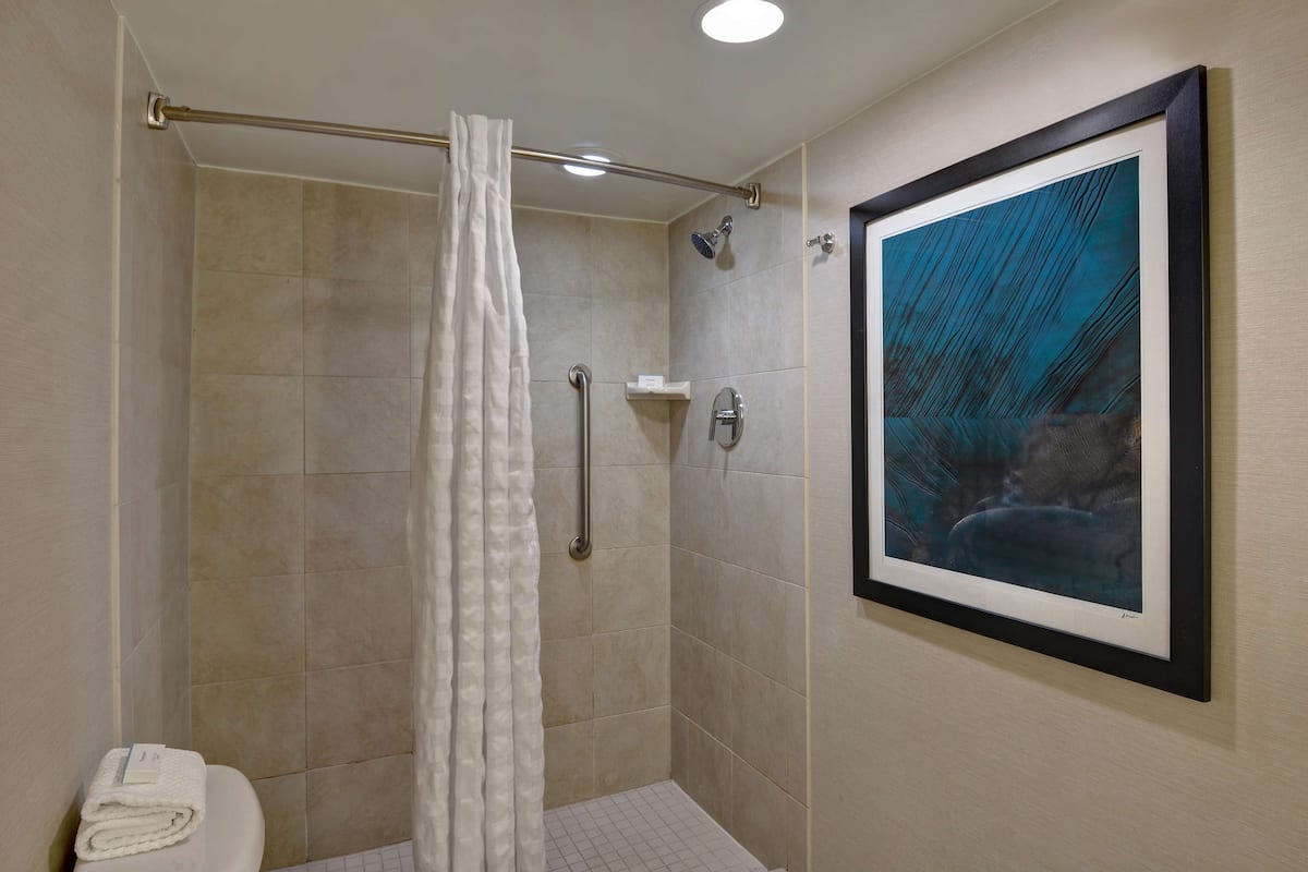 A shower with a white curtain, a framed artwork on the wall, and a white towel on the floor.