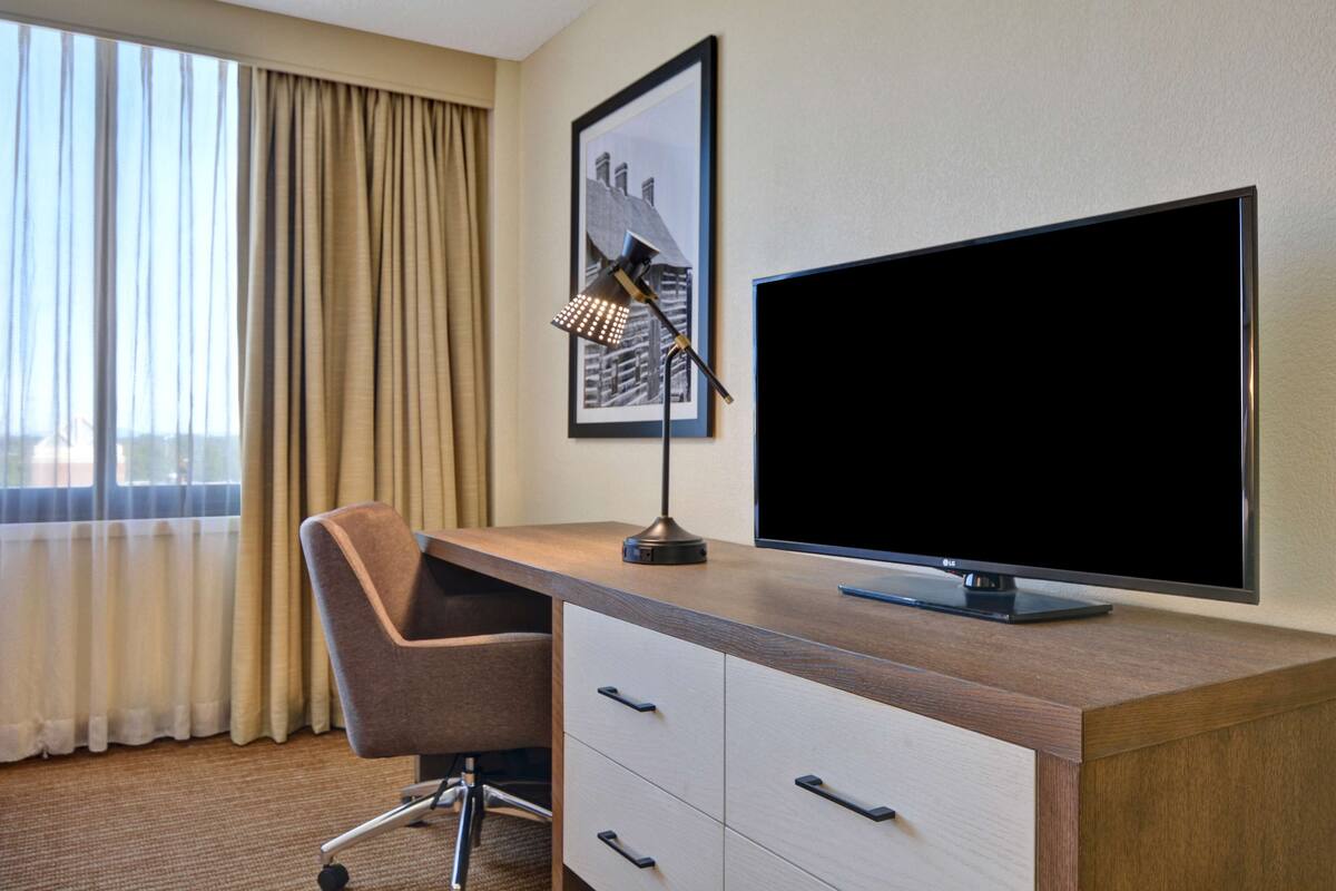 A hotel room with a desk, a chair, a flat-screen TV, a framed picture on the wall, and a window with curtains.