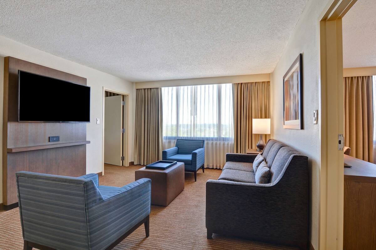 A hotel room with a flat-screen TV, a sofa, a chair, a coffee table, and a desk.