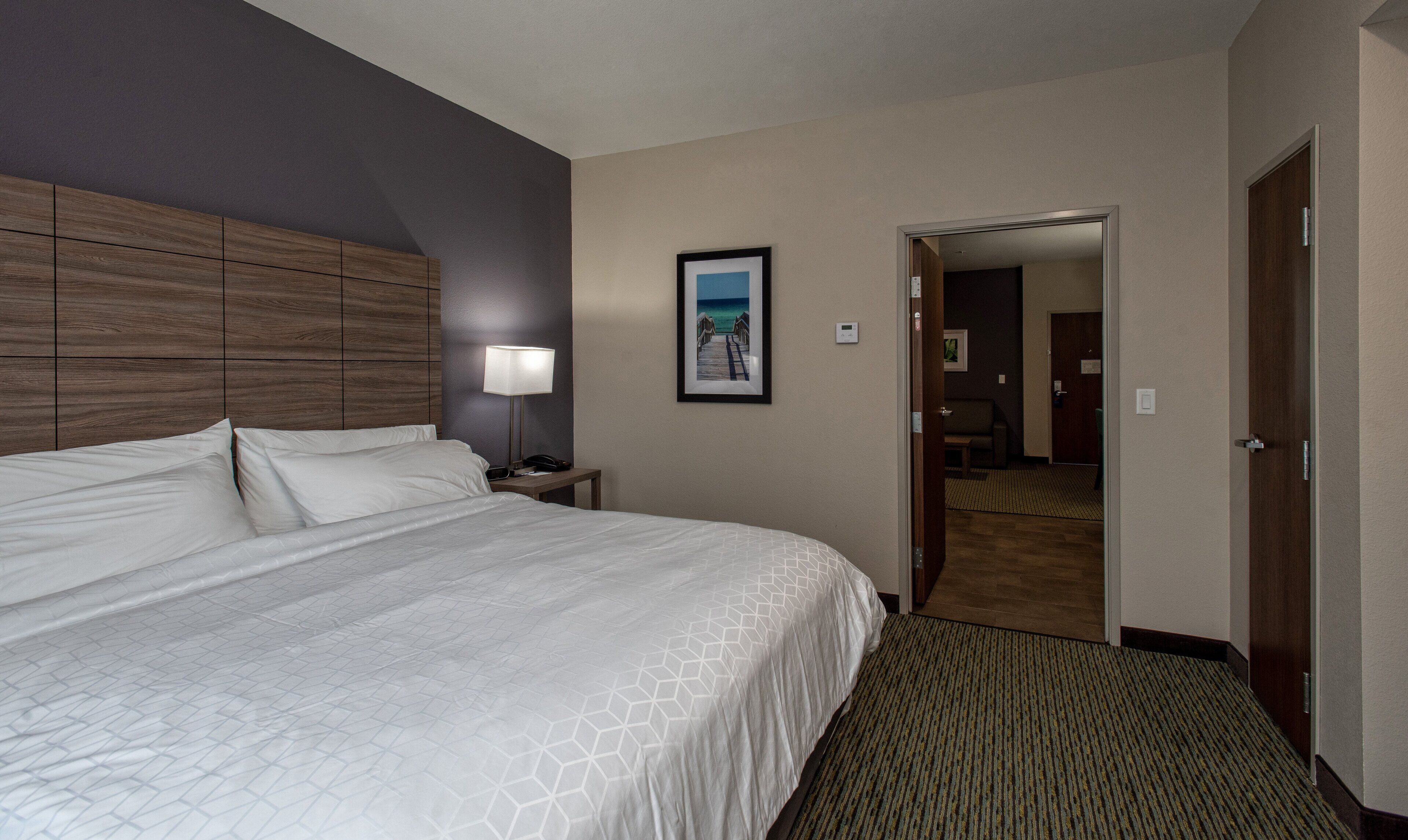 A hotel room with a large bed, a nightstand with a lamp, a framed picture on the wall, and a door leading to another room.