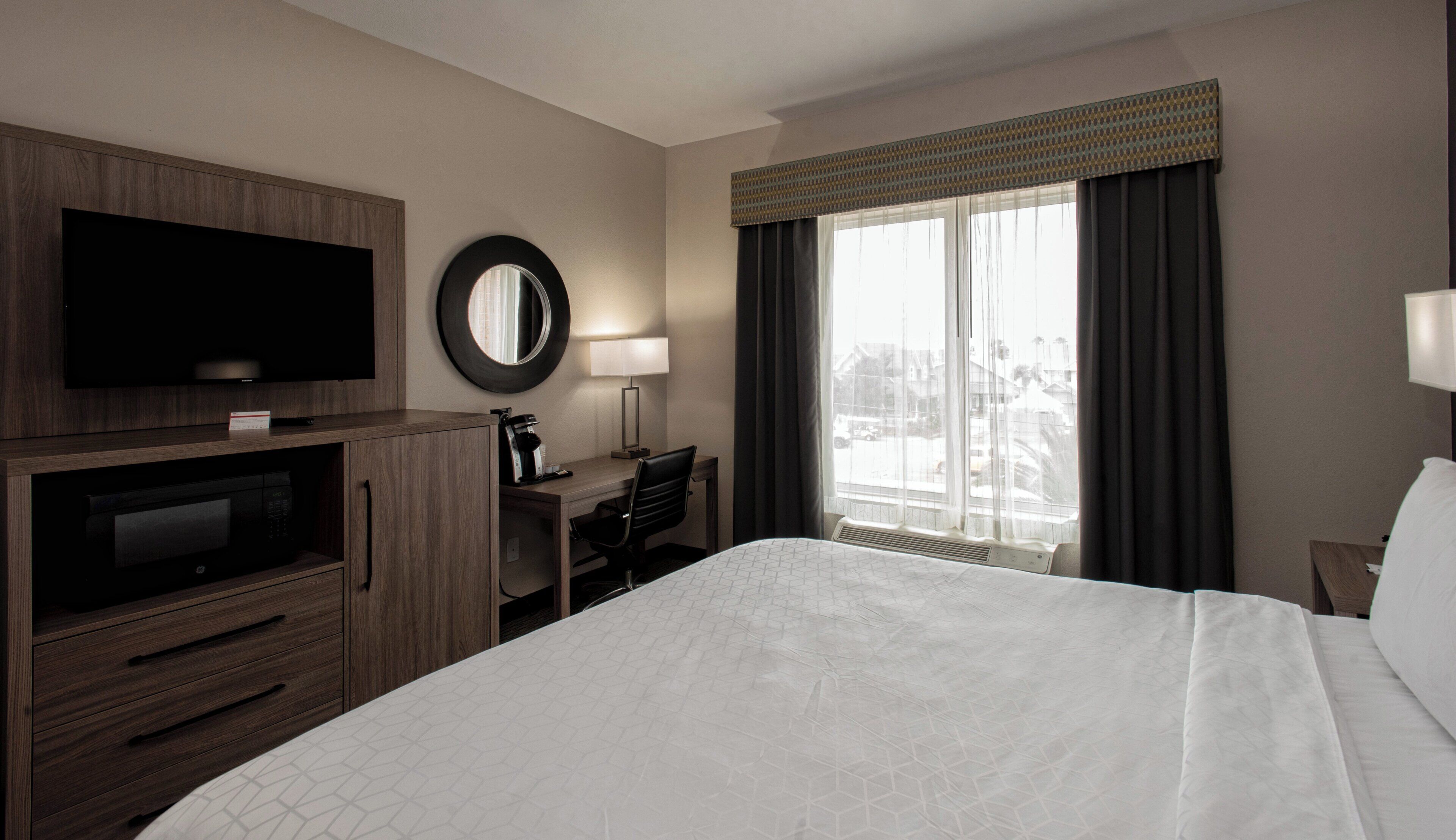 Une chambre d’hôtel équipée d’un lit, d’un bureau, d’une télévision, d’un miroir et d’une fenêtre avec des rideaux.
