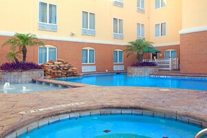 A swimming pool with a stone deck, surrounded by a building with multiple windows and a brick facade.