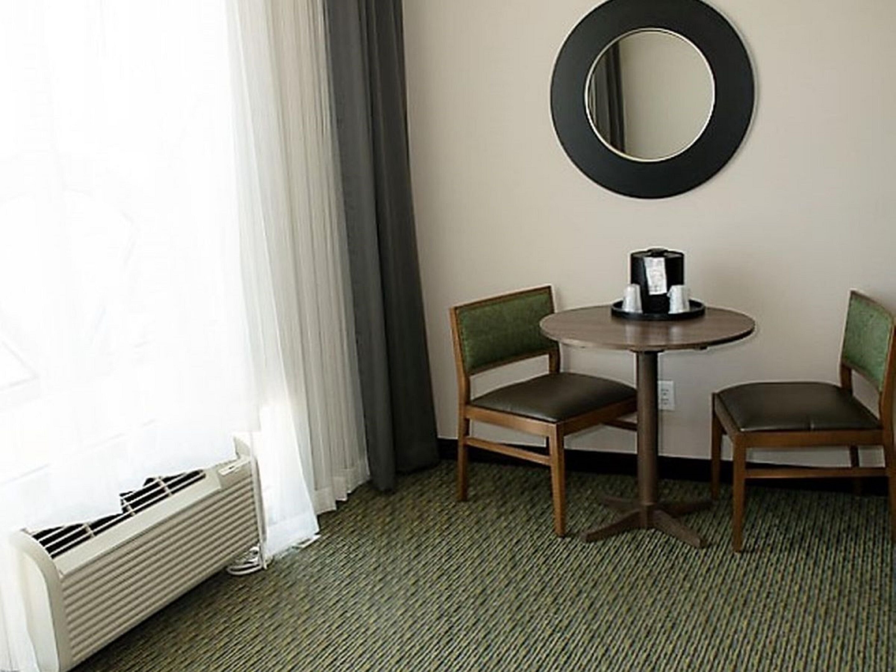 A hotel room with a round table, two chairs, a mirror, a radiator, and a window with curtains.