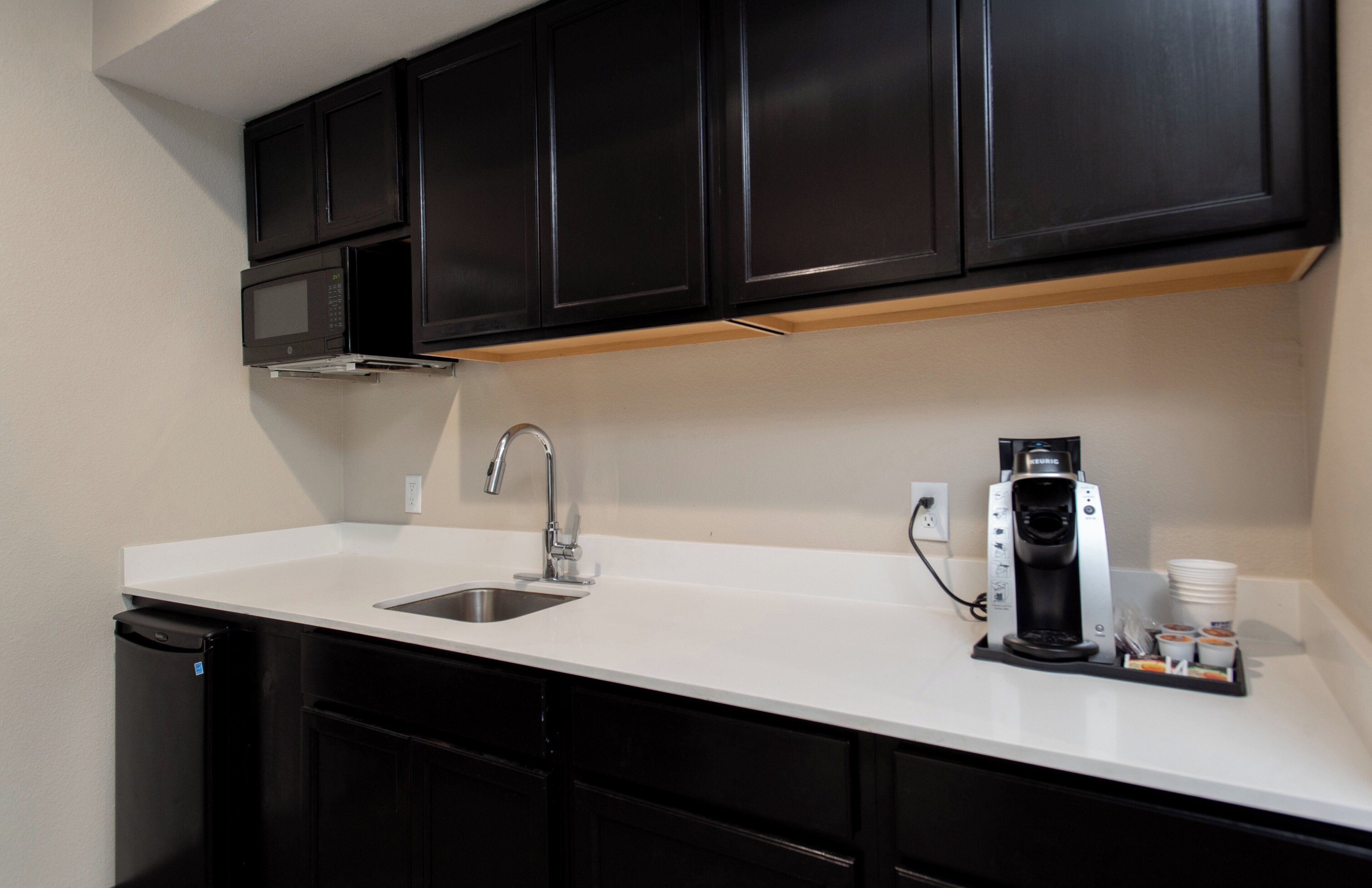 A kitchen with a white countertop, a stainless steel sink, black cabinets, a microwave, and a coffee maker.