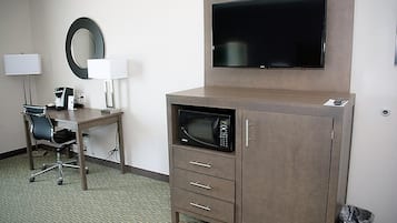 A hotel room with a desk, chair, lamp, television, microwave, and a trash bin.