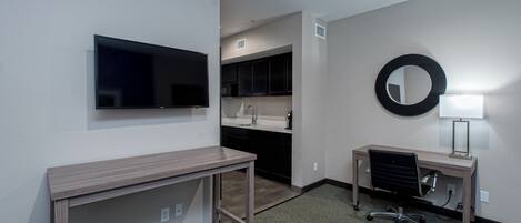 Une chambre d’hôtel équipée d’un téléviseur à écran plat, d’un bureau avec une chaise, d’un petit miroir et d’une kitchenette comprenant un évier et des placards.