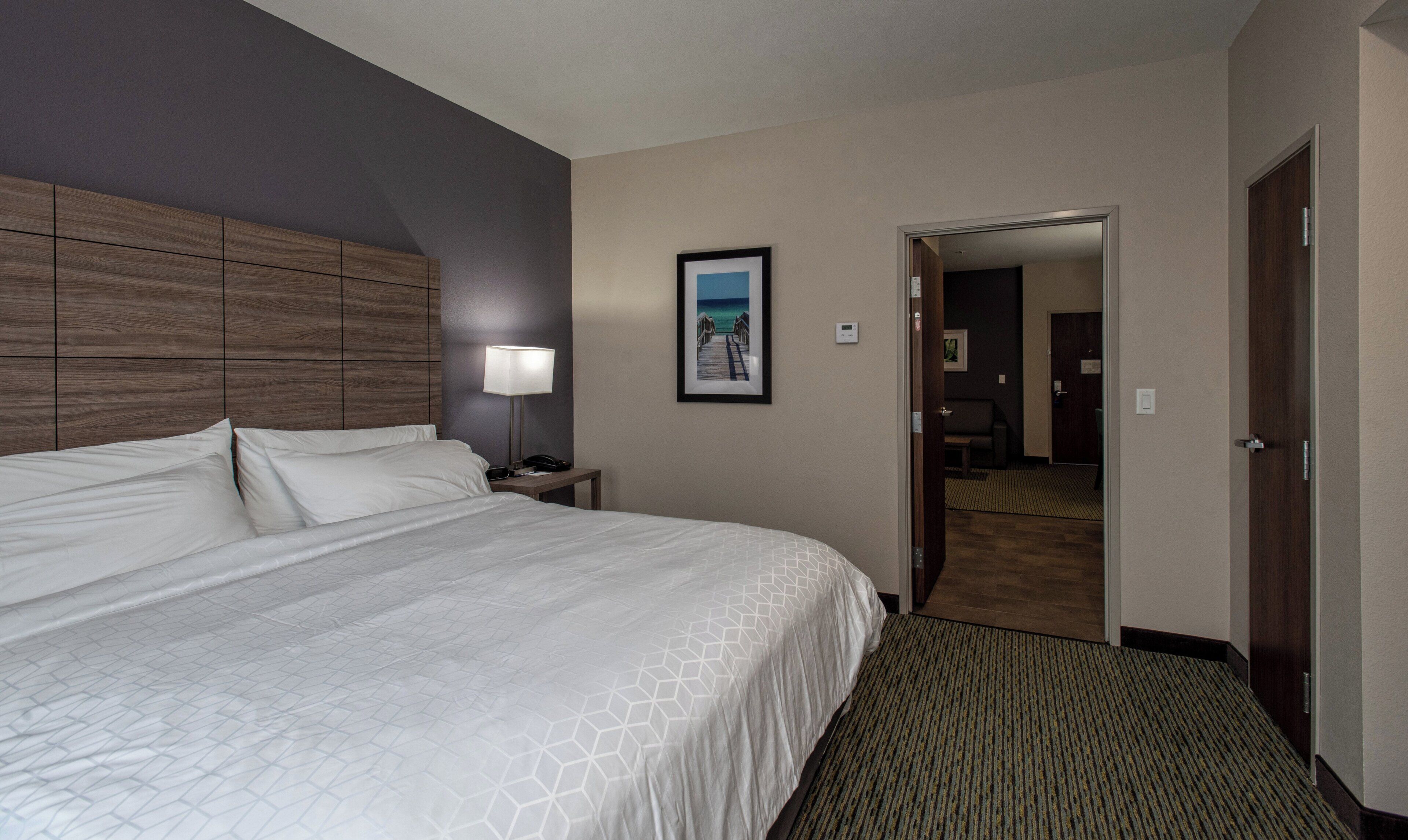 A hotel room with a large bed, a nightstand with a lamp, a framed picture on the wall, and a door leading to another room.