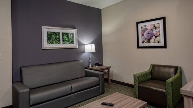 A hotel room with a grey sofa, a green armchair, a small wooden table, a lamp, and two framed pictures on the wall.