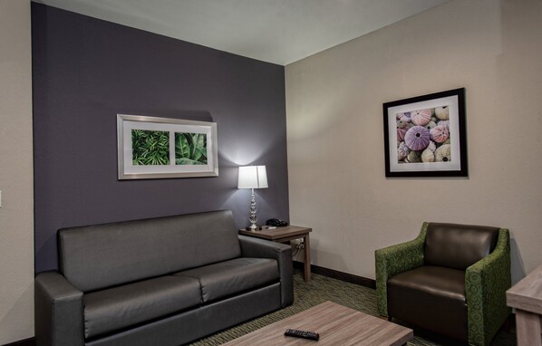 Habitación de hotel con un sofá gris, un sillón verde, una pequeña mesa de madera, una lámpara y dos cuadros en la pared.
