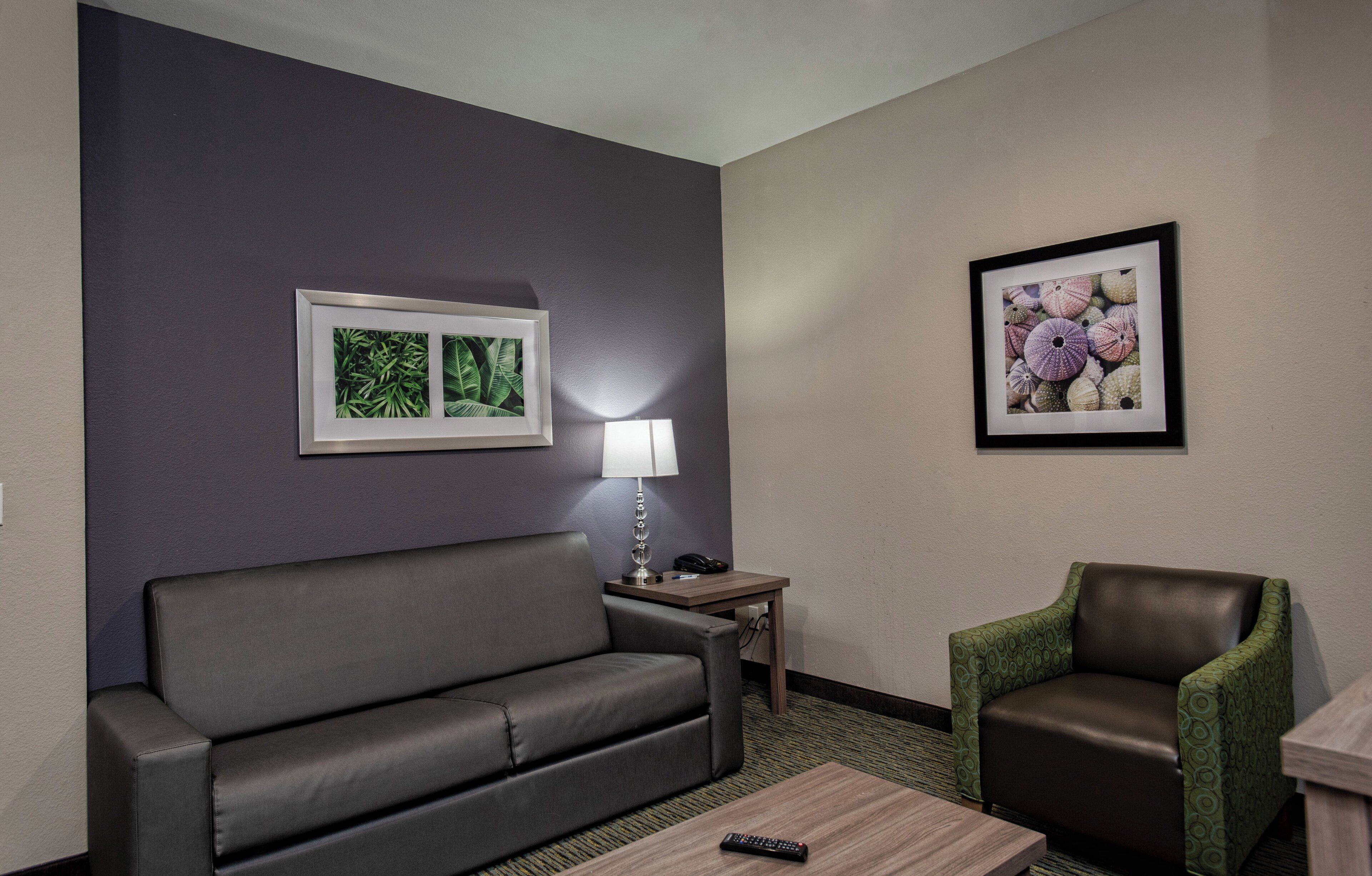 Habitación de hotel con un sofá gris, una mesita con una lámpara, un sillón verde y dos cuadros en la pared.