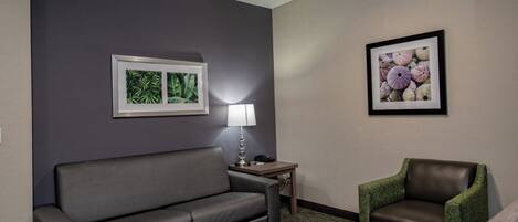 Habitación de hotel con un sofá gris, una mesita con una lámpara, un sillón verde y dos cuadros en la pared.