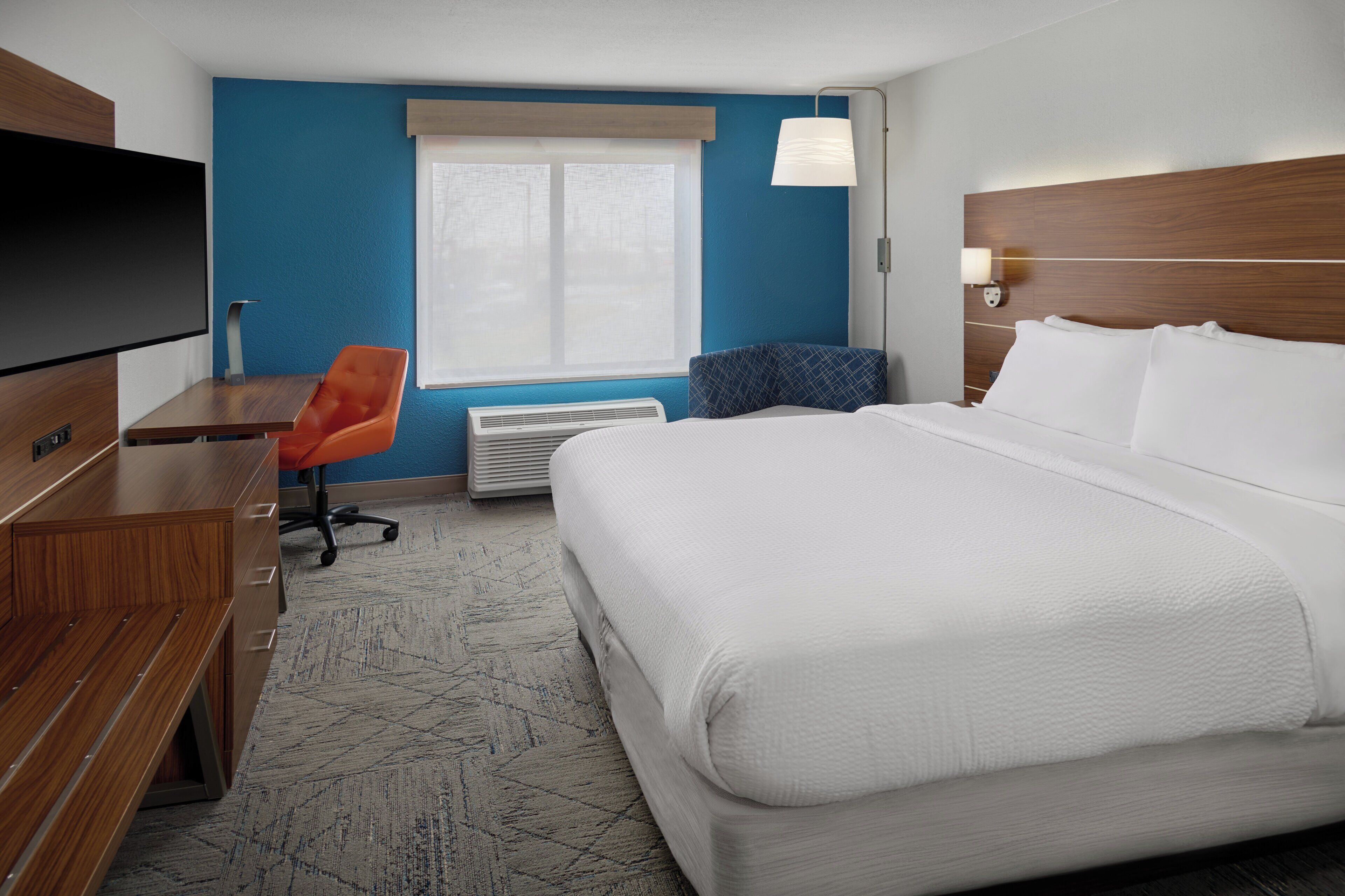Habitación de hotel con cama, escritorio, silla naranja, televisor de pantalla plana y ventana con persianas.