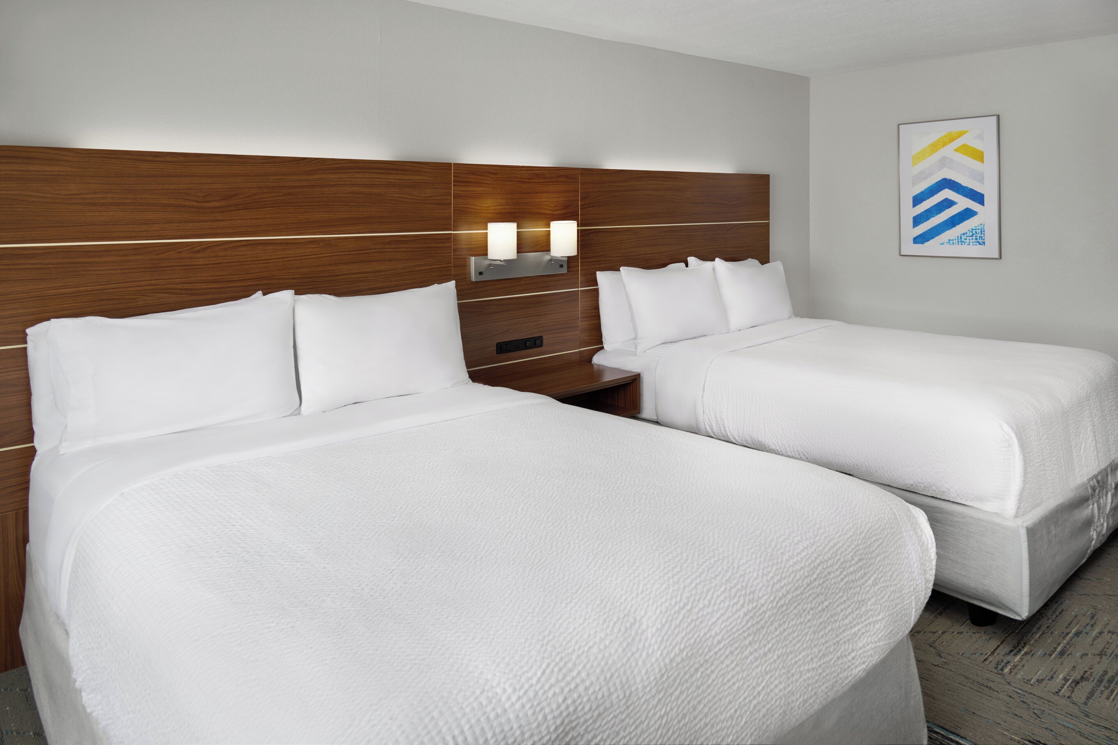 Habitación de hotel con dos camas, cabecero de madera, lámparas de noche e imagen enmarcada en la pared.