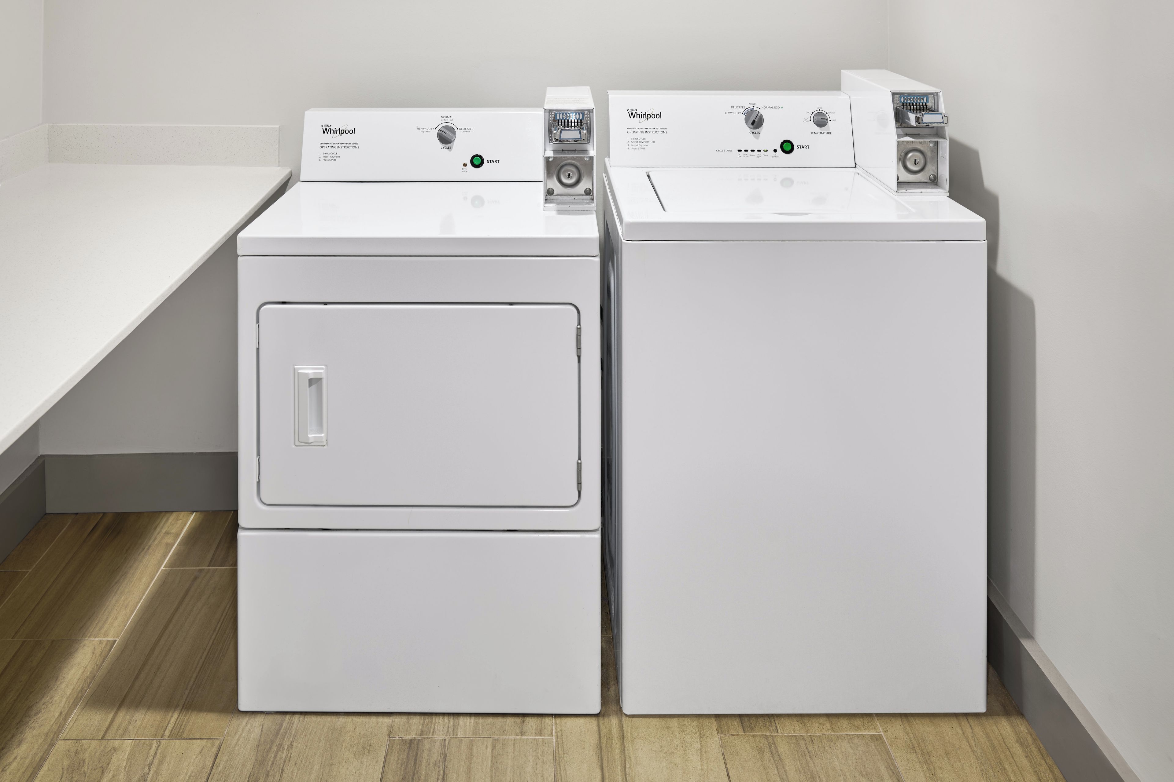 Dos lavadoras Whirlpool blancas de carga frontal en un cuarto de lavado con piso de madera y encimera blanca.
