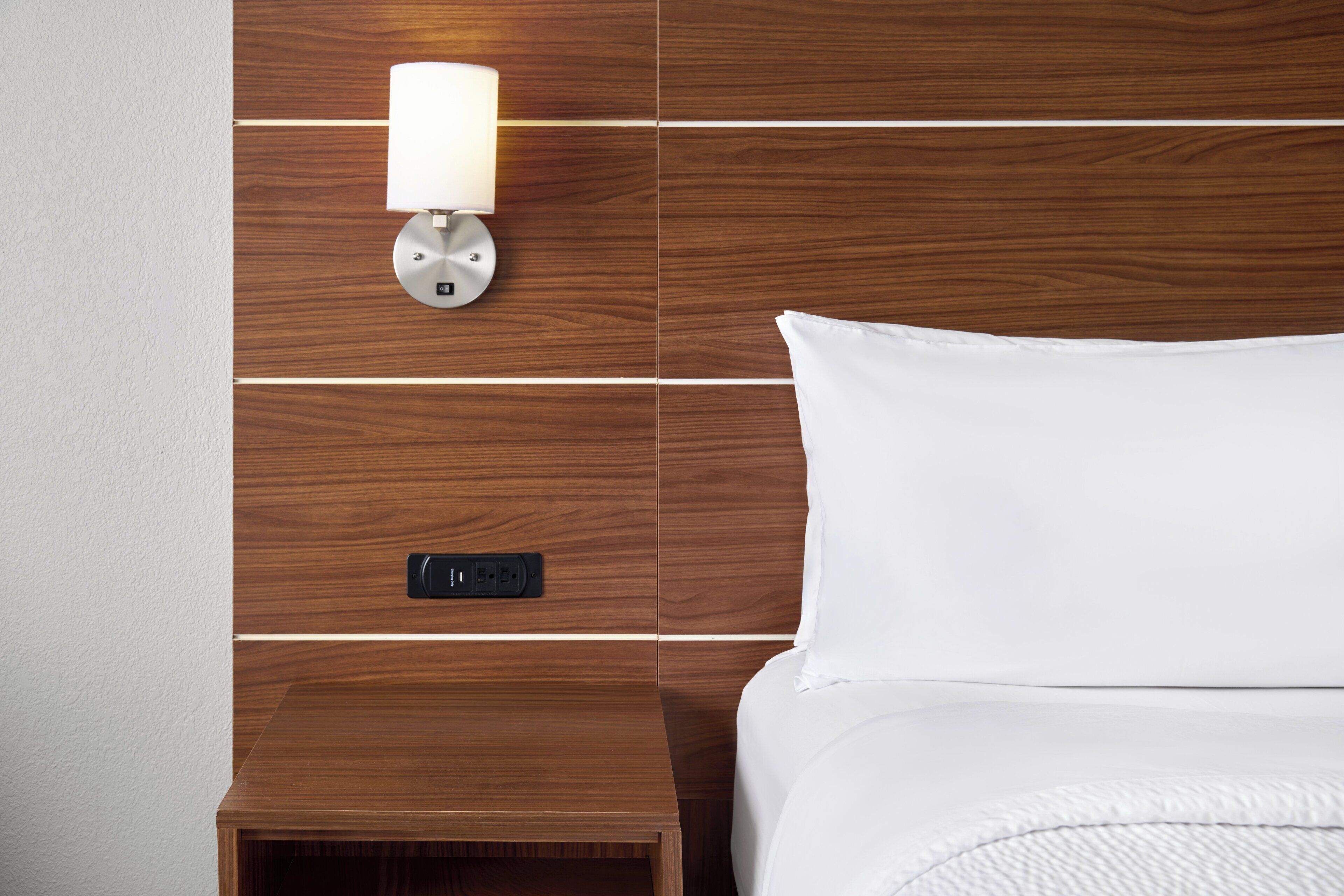 A neatly made bed with white linens, a wall-mounted lamp, and a power outlet on a wooden panel wall.