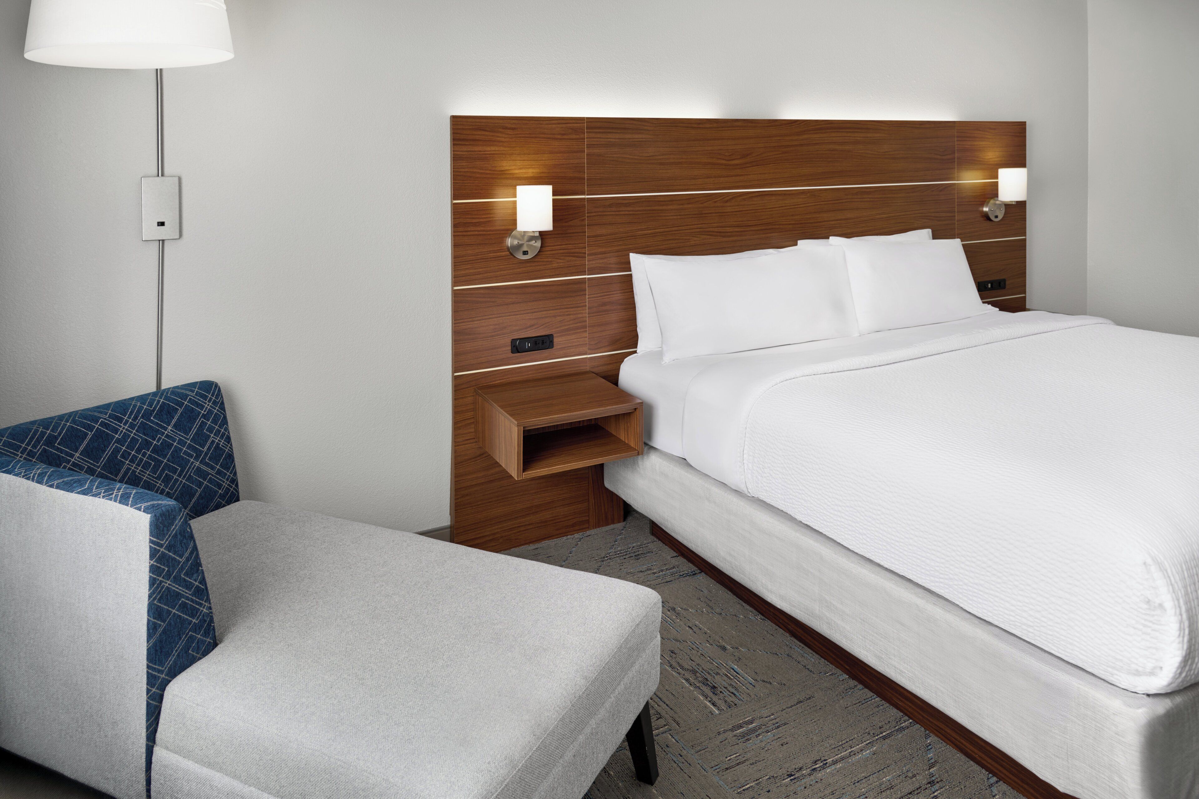 A hotel room with a wooden headboard, a bed with white bedding, a nightstand, a lamp, and a chair with a blue cushion.