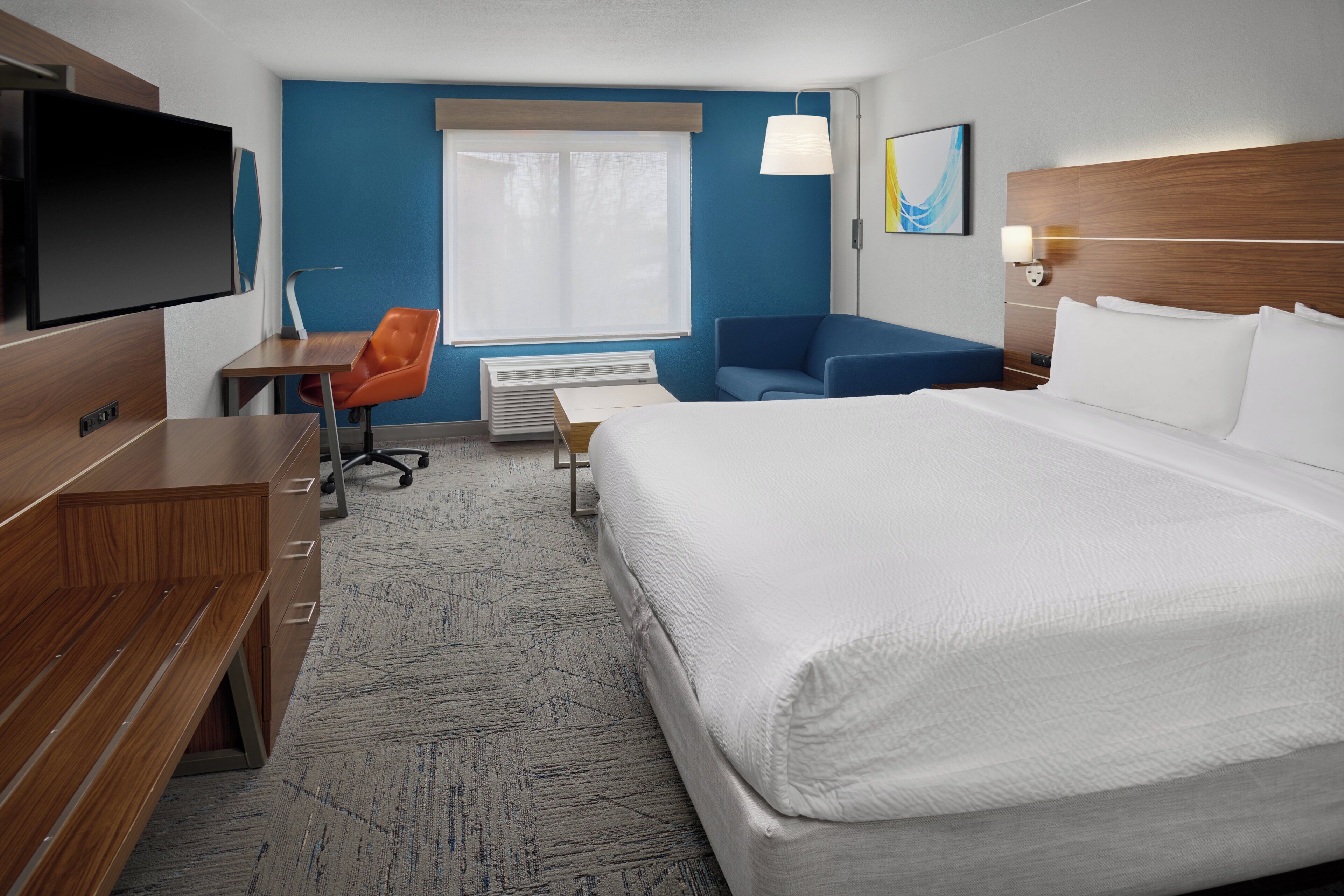 Une chambre d’hôtel avec un grand lit, un bureau avec une chaise, une télévision et une fenêtre avec des stores.