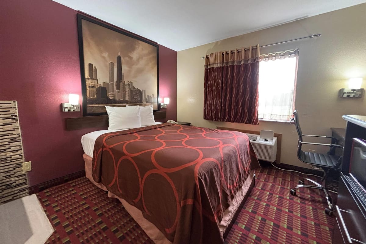 Quarto de hotel com uma cama, uma escrivaninha com um laptop, uma janela com cortinas e uma pintura de paisagem urbana na parede.