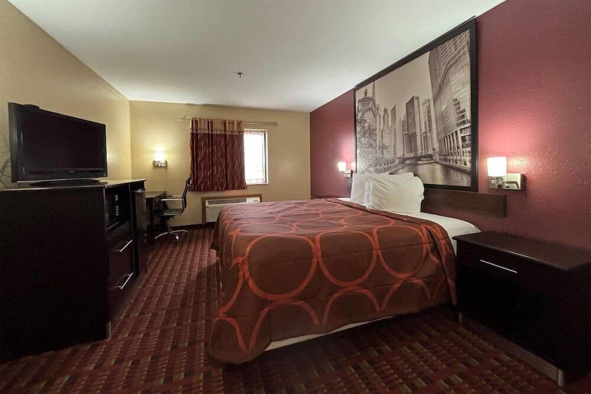 Quarto de hotel com cama, televisão, escrivaninha, cadeira, janela com cortinas e uma pintura de paisagem urbana na parede.