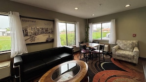 A hotel room with a black leather sofa, a wooden coffee table, a patterned armchair, a dining table with chairs, and large windows with curtains.