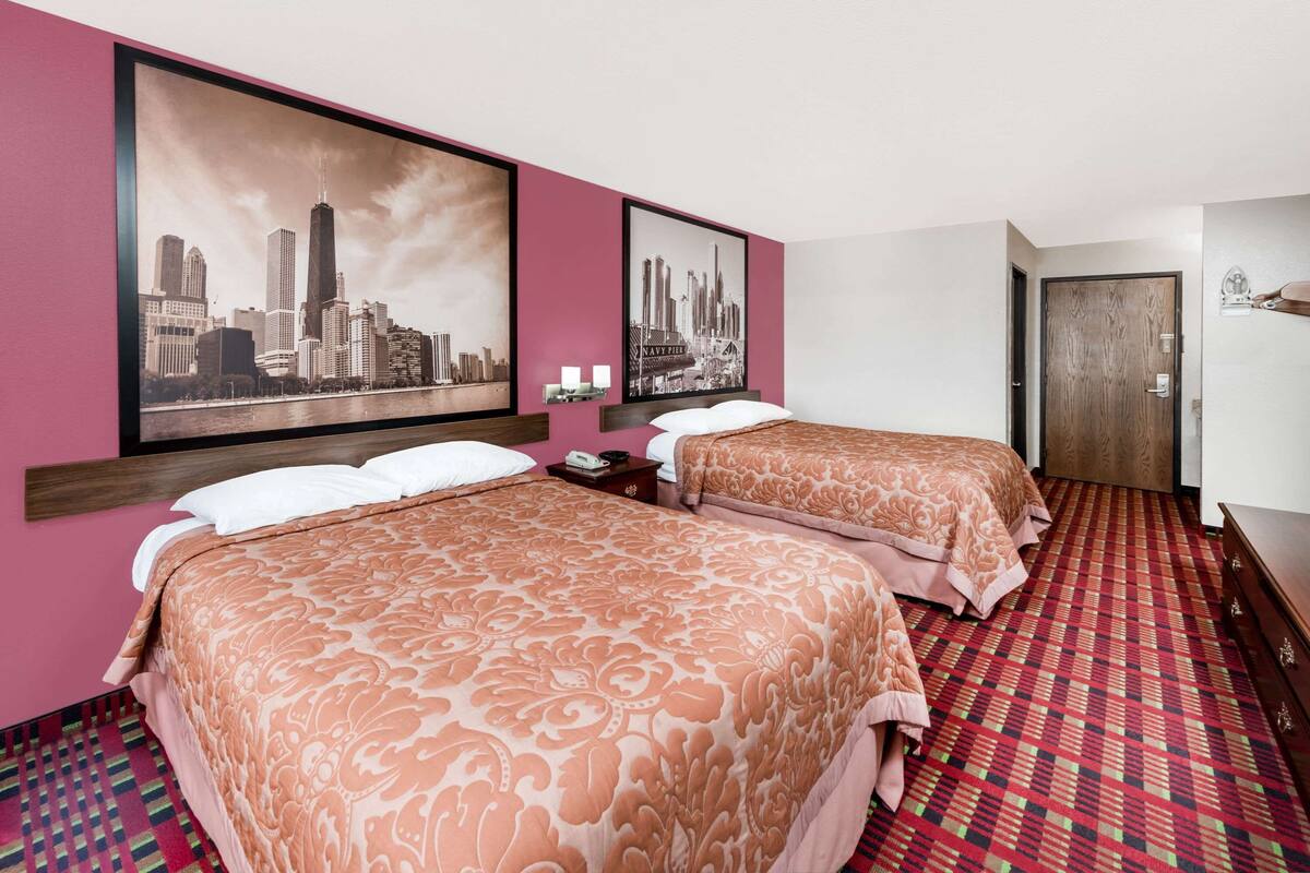 Quarto de hotel com duas camas, uma pintura urbana na parede, tapete vermelho estampado e porta de madeira.