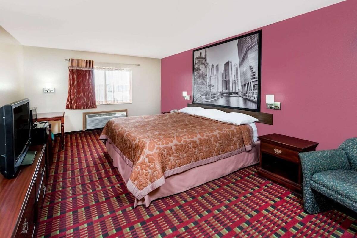Quarto de hotel com cama, televisão, janela com cortinas, quadro na parede e carpete estampado.