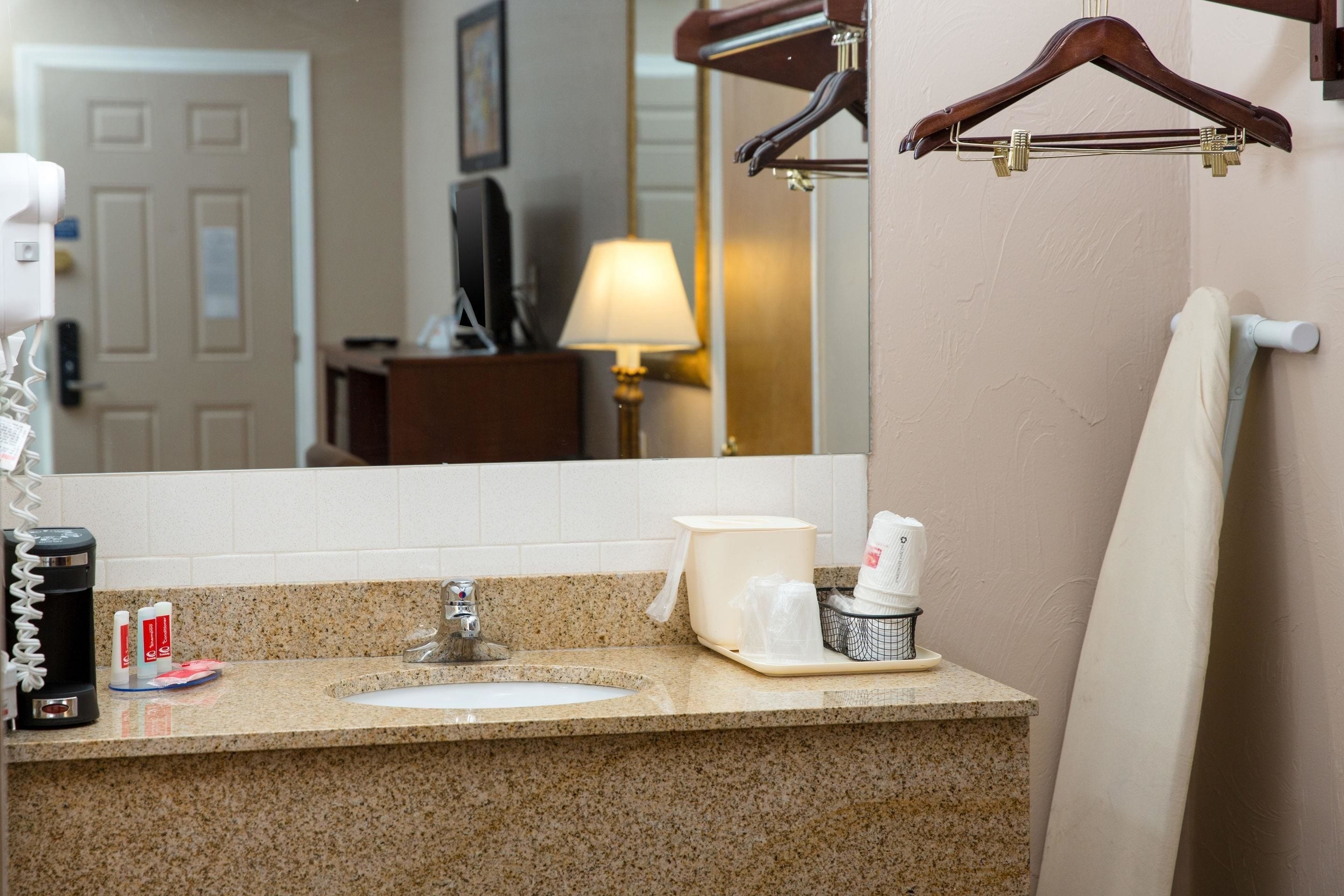 Un baño de hotel con encimera de granito, lavamanos, espejo, toallero y cafetera.