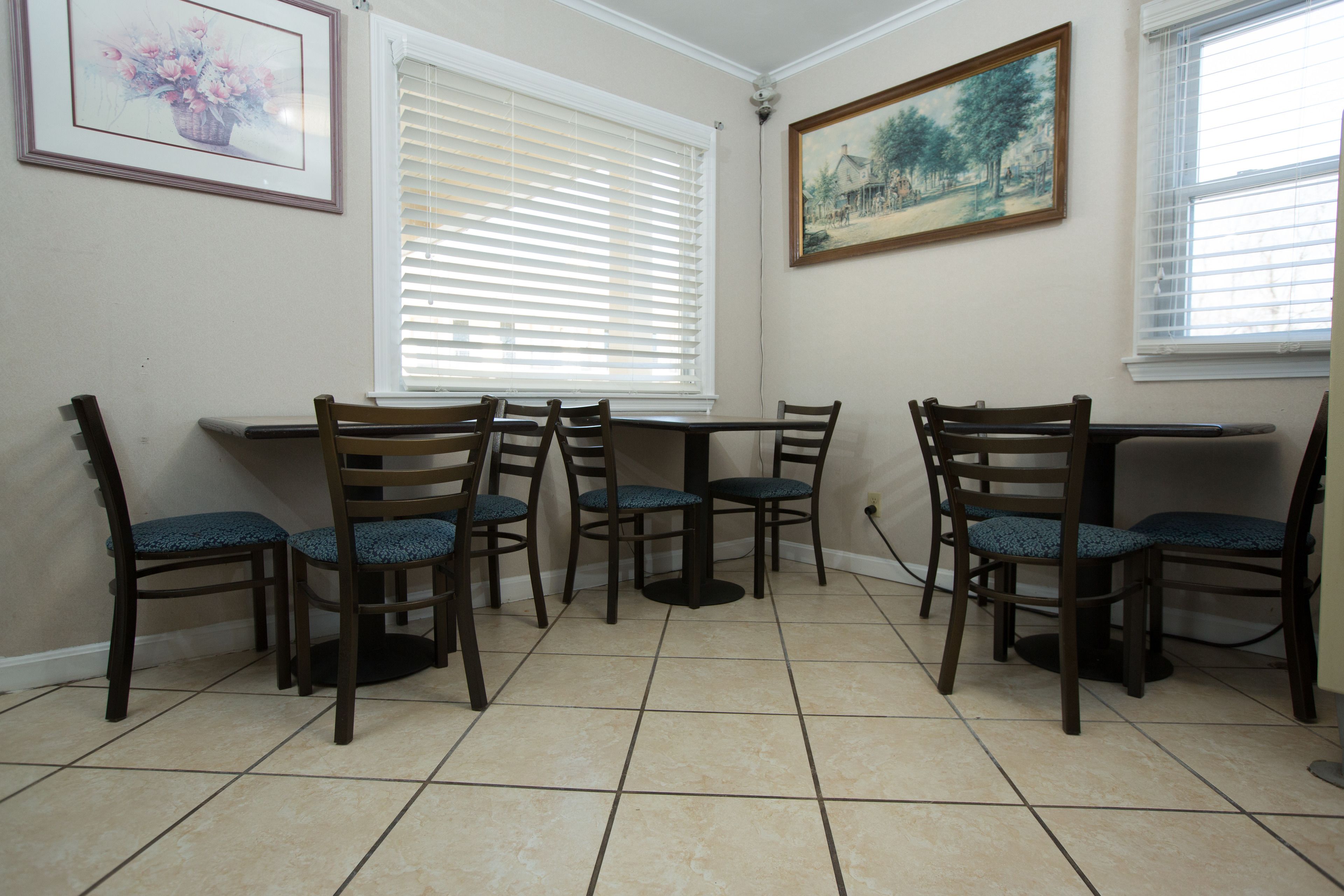 A dining area with six wooden chairs and a table, white window blinds, and two framed artworks on the walls.