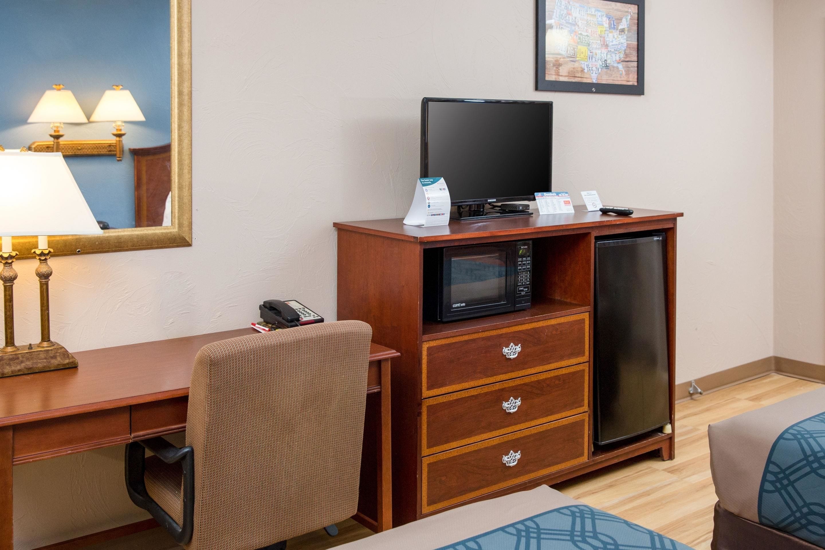 A hotel room with a wooden desk, a chair, a television, a microwave, a refrigerator, and a bed with a patterned cover.