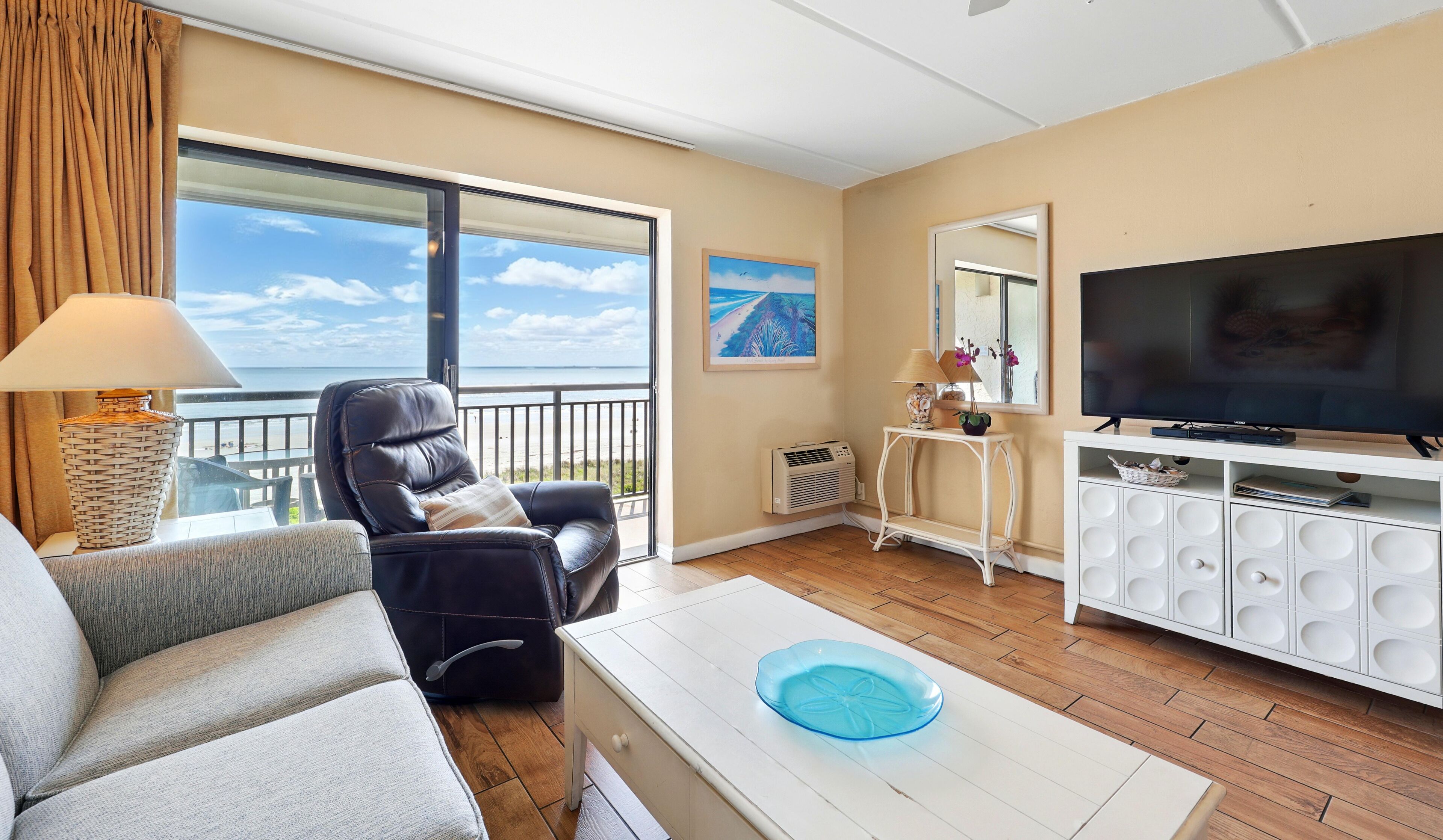 A living room with a leather sofa, a white coffee table with a blue glass bowl, a TV, and a large window with a view of the beach.