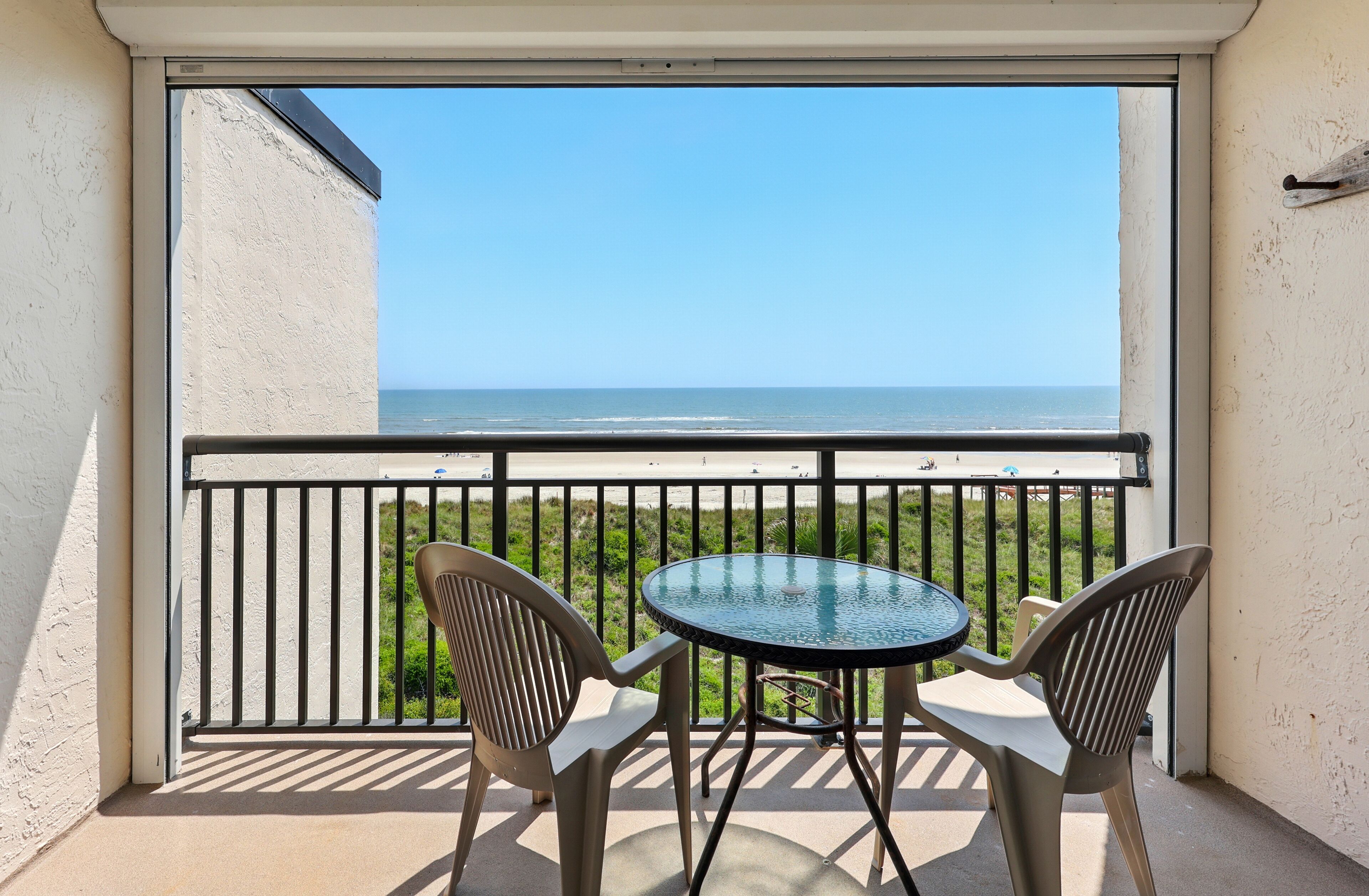 A balcony with a glass table and two chairs, overlooking a beach with the ocean and a clear sky.