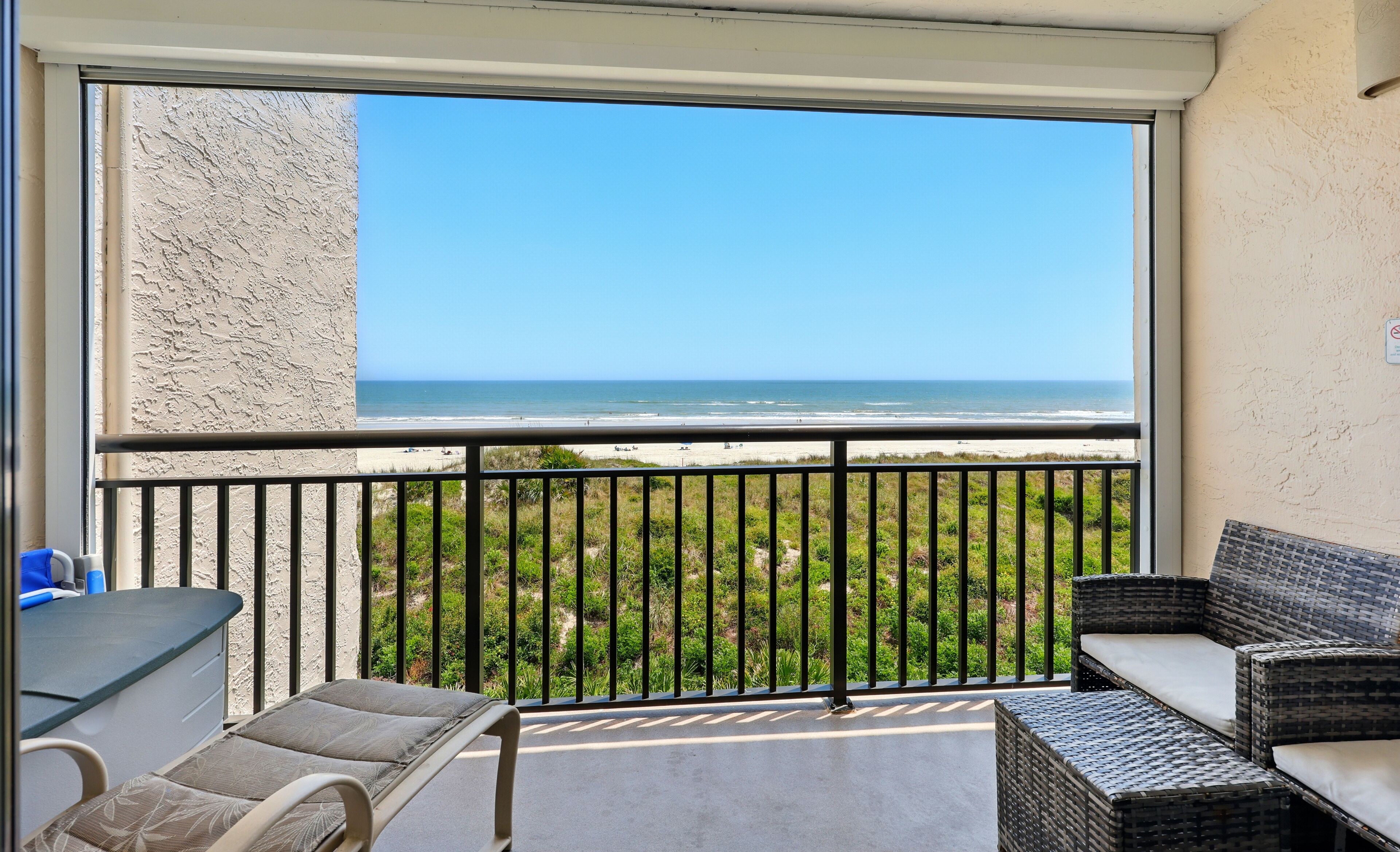A balcony with a view of the beach, a wicker sofa, and a table with chairs.