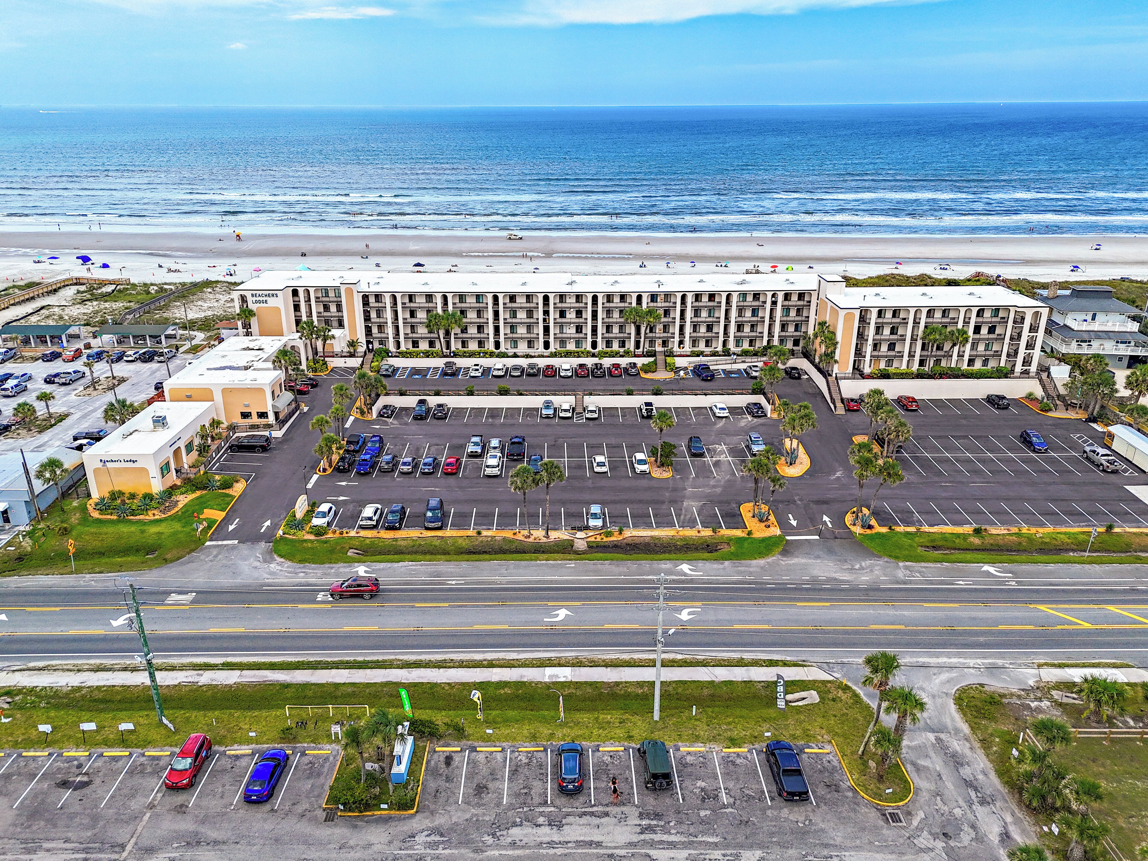 Ein großer Parkplatz mit mehreren Reihen Autos, ein Hotelgebäude mit Blick auf den Strand und ein Strand im Hintergrund.