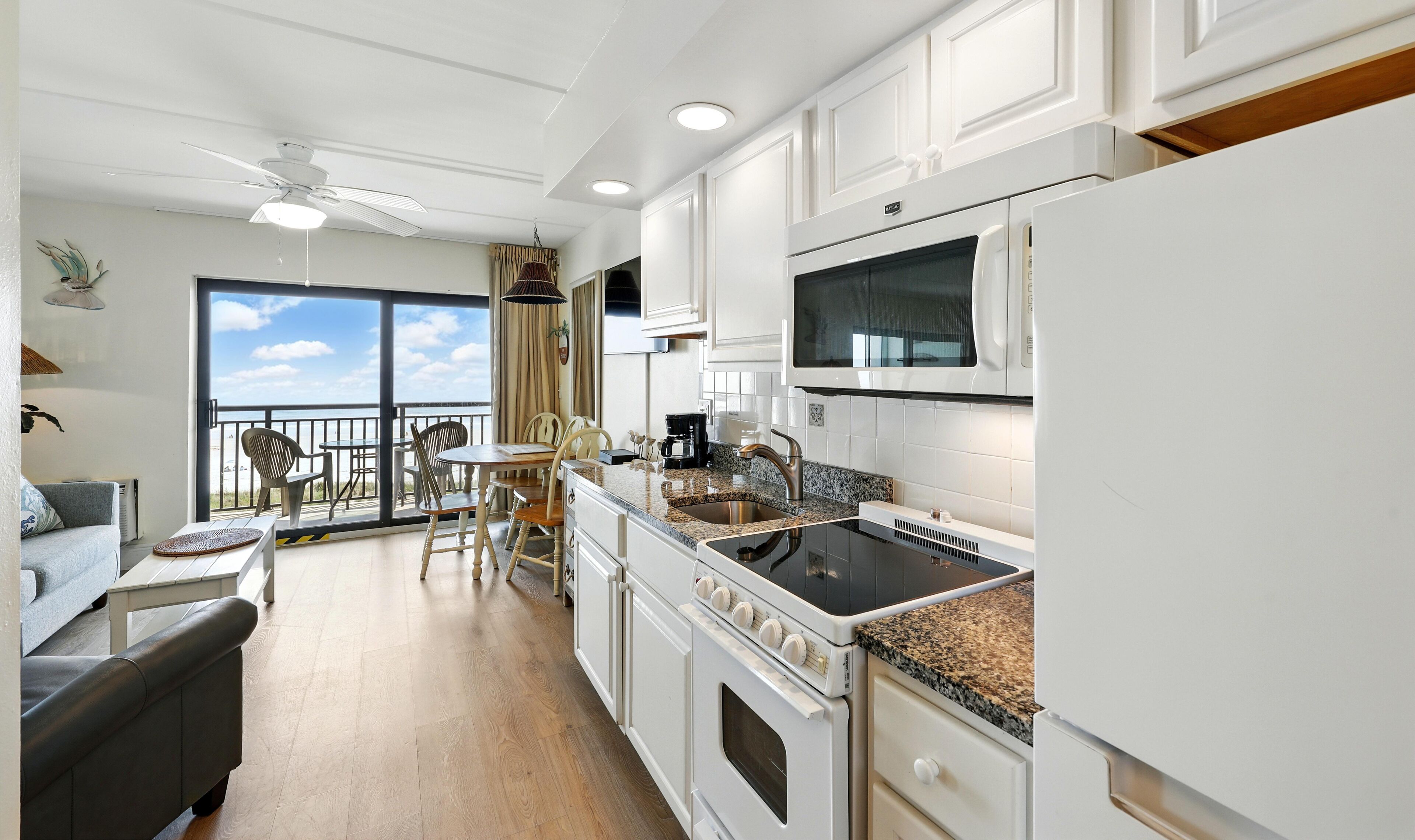 A modern kitchen with white cabinets, a built-in microwave, and a stove. There is a dining area with a table and chairs, and a balcony with a view.