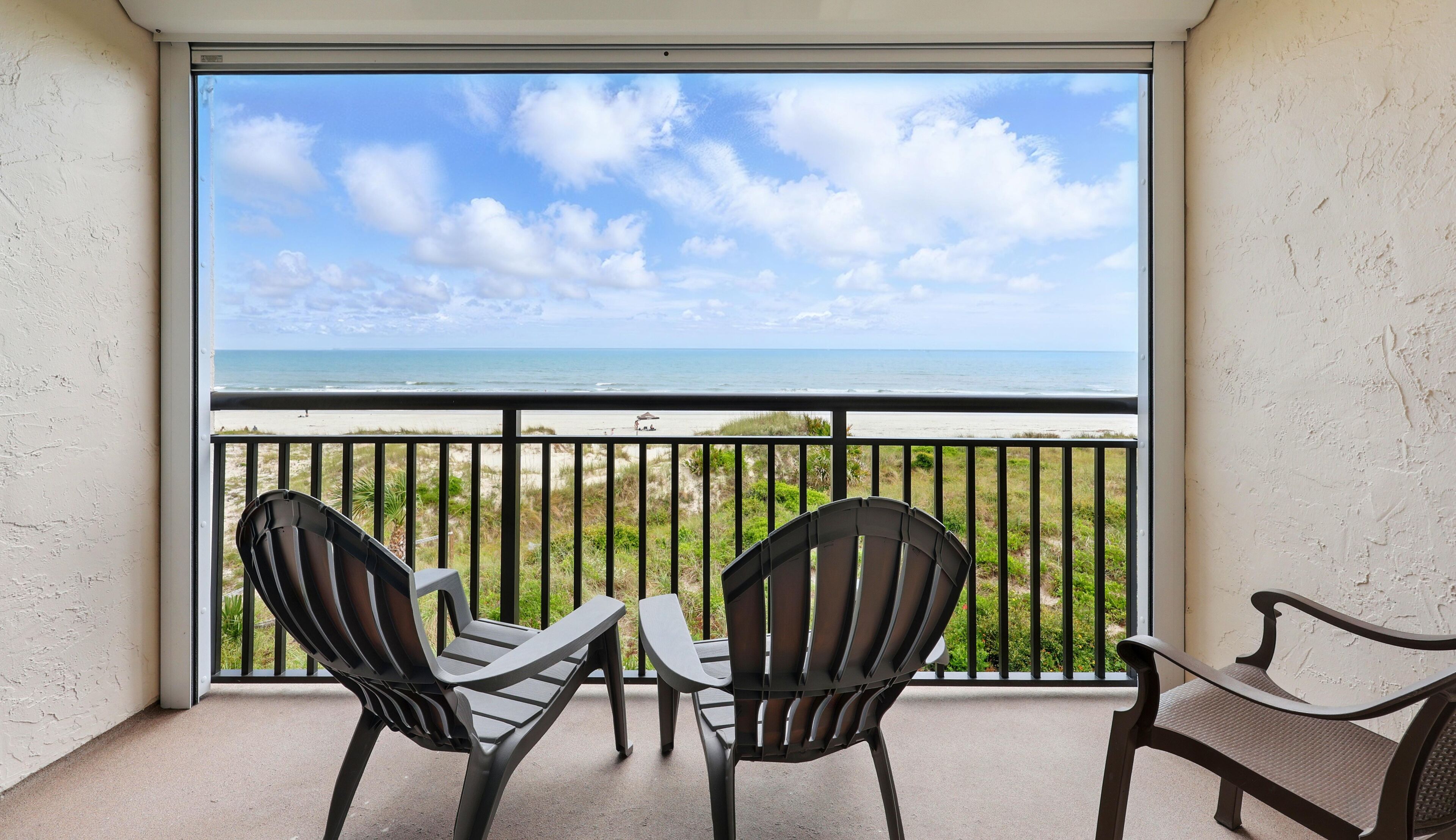 Un balcon avec deux chaises et une petite table, donnant sur une plage sous un ciel dégagé.
