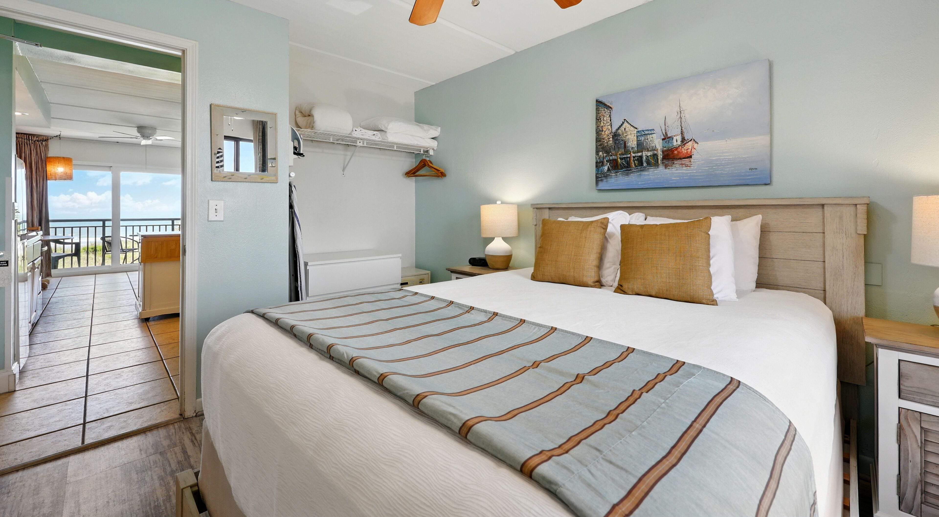 A hotel room with a large bed, a painting of a coastal scene, a balcony with a view, and a ceiling fan.
