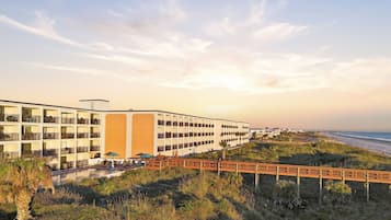 Un hotel frente a la playa con varios balcones, una piscina y un paseo marítimo de madera que lleva a la playa.