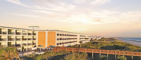A beachfront hotel with multiple balconies, a pool, and a wooden boardwalk leading to the beach.