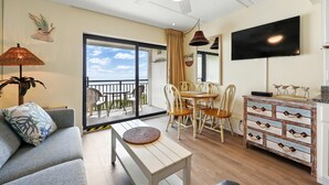 Una sala de estar con sofá, una mesa de centro, televisor, una mesa de comedor con sillas, un balcón con vistas y un adorno en la pared.